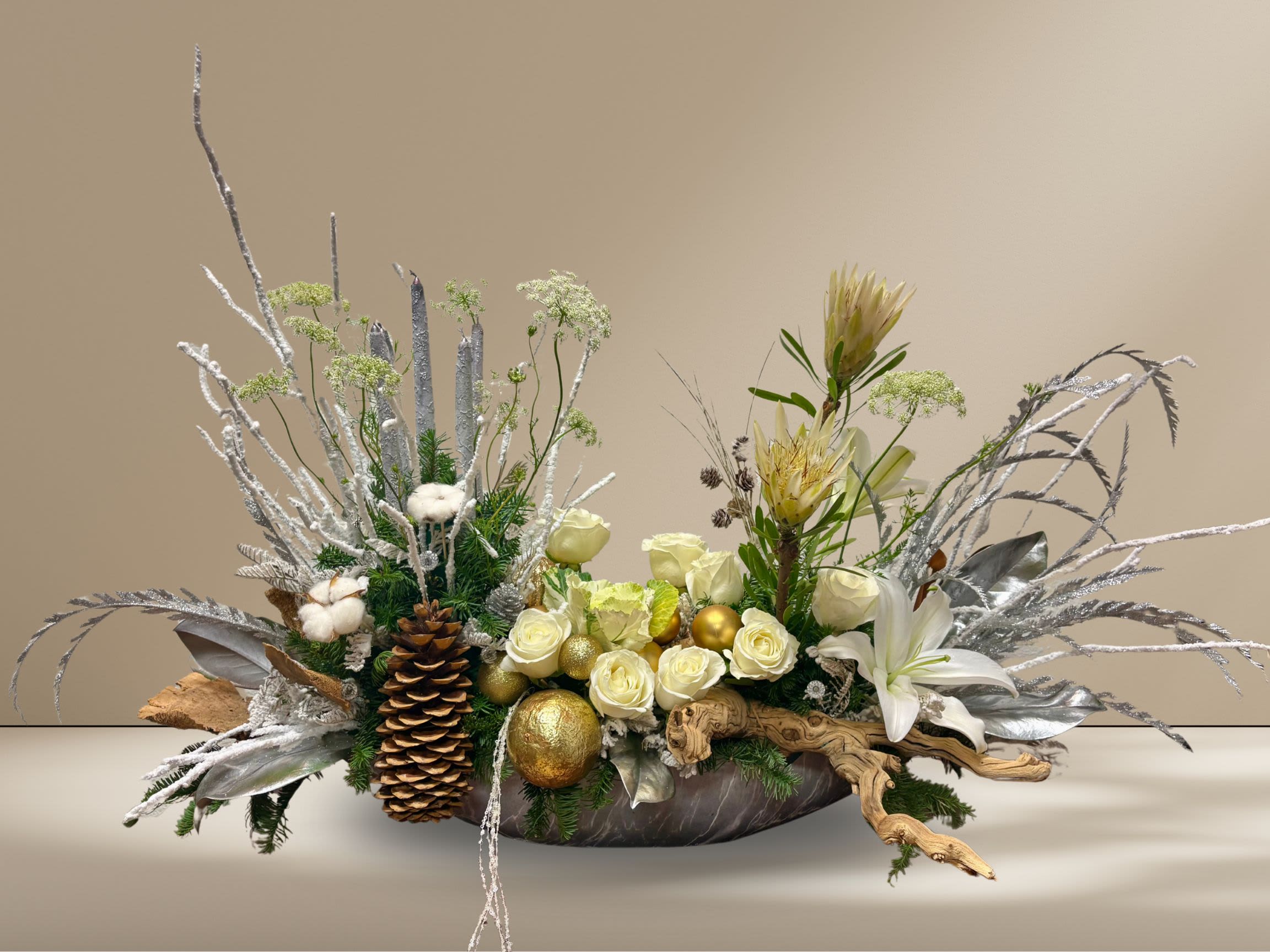 Grand Winter Majesty - A grand holiday centerpiece that brings the magic of a winter forest indoors. Designed in a long marble vase, Winter Majesty blends natural elements—pinecones, maple burl wood, driftwood, silver-painted magnolia leaves, and pine greenery—with an elegant mix of white roses, kale, protea, and lilies. Frosted branches and shimmering silver accents add a touch of winter sparkle, while sculptural branches and candles create graceful, elevated height on one side. A luxurious, artful statement piece for any festive table or celebration.
