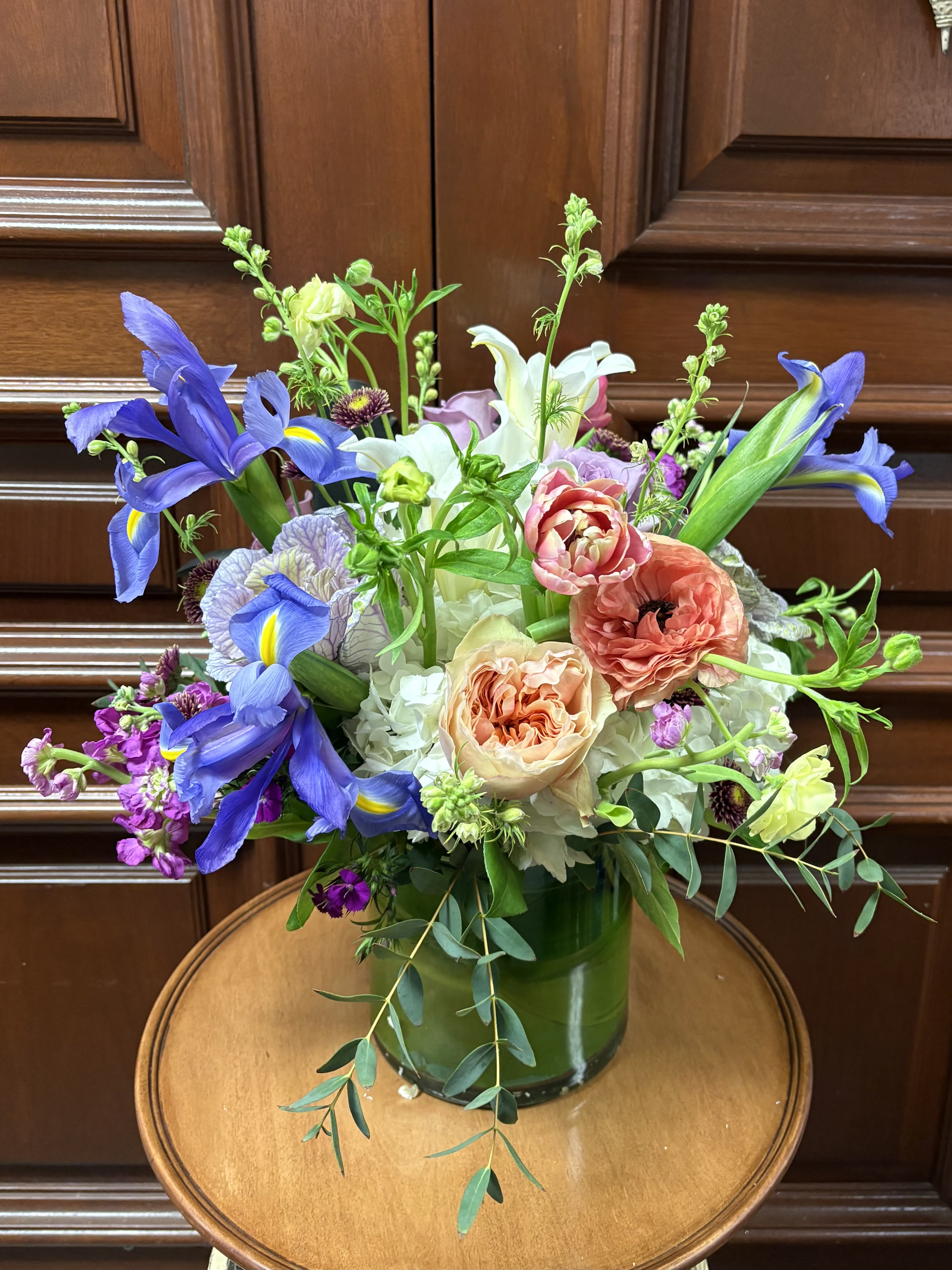 Spring Garden Meadow - This Spring Garden has soft color garden roses (depending on availability), Kale, Seasonal Ranunculus, Bright Blue Iris, Lilies, Hydrangaes, and Stock in a grand round crystal vase.
