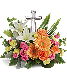 Low arrangement of white lilies, orange gerbera daisies, roses and pink spray roses around a silver cross