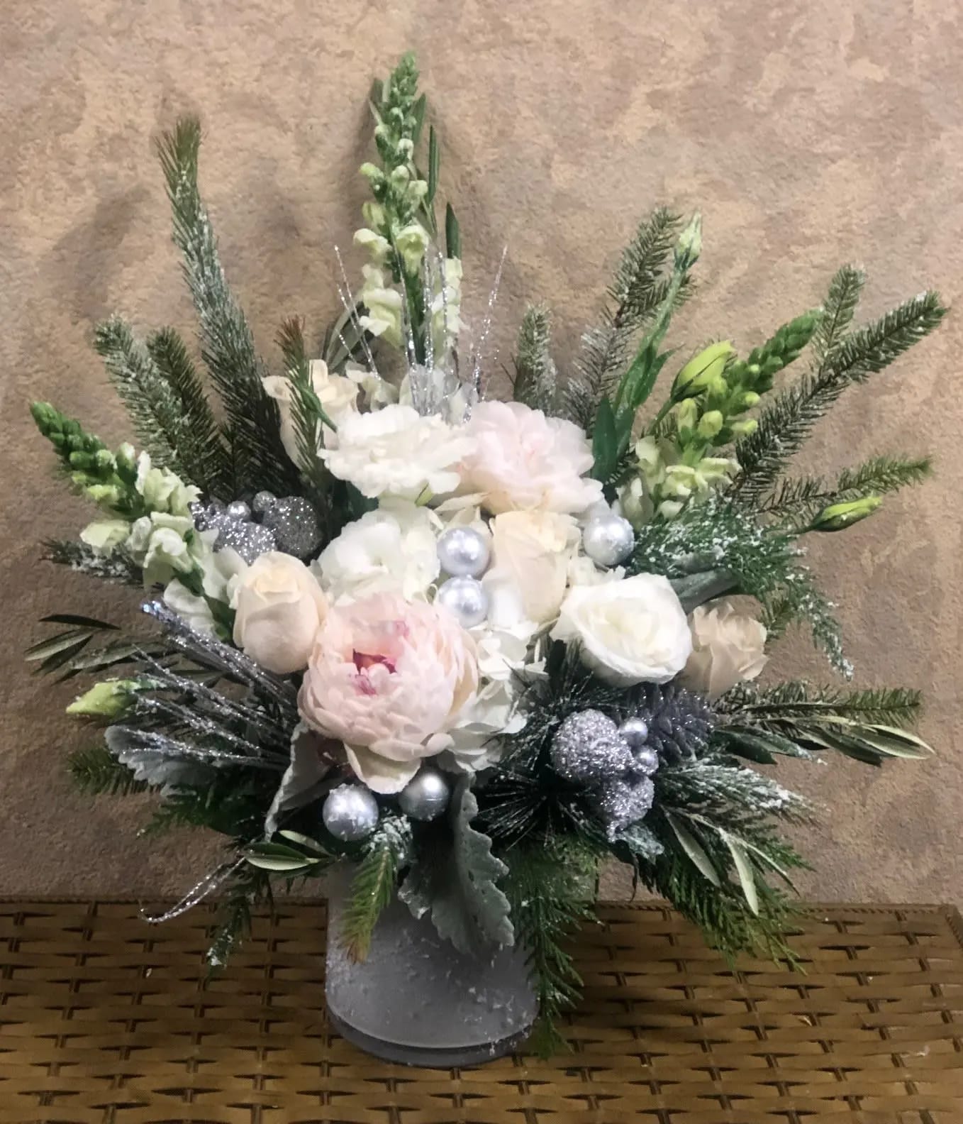 Have a Glistening Holiday - Type of Flowers: White Snapdragons, White Snapdragon, White Lisianthus, White Roses, White Peonies, Silver Pine greens, Dusty Miller and silver branches, pine cones, band silver ornaments in a short, white, crystal vase. Availability: All year round Substitute Available: Yes Design View: Front Facing View Photo shown: Regular 