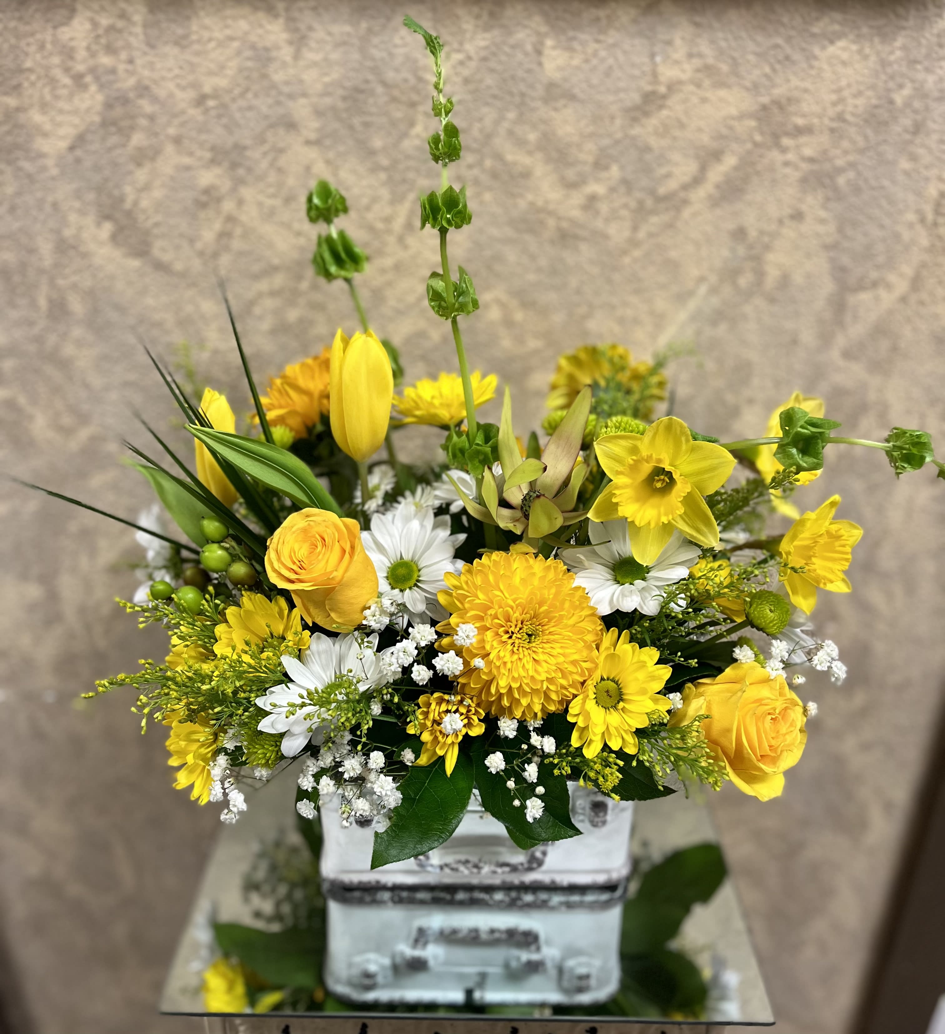 Glory - Recipe: Yellow Roses, White Daisies, Gerbera Daisies, Tulips, Button Mums, Chrysthemums, Bells of Ireland, hypericum Berries, Babies Breath in a suitcase vase. Availability: All year round Design View: Symmetric Front Facing View Photo shown: Standard 