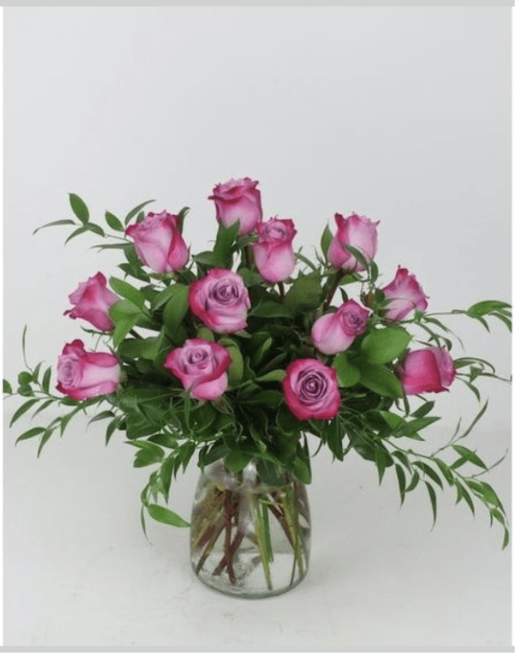 PURPLE DREAM ROSES - A loose, rounded cluster of purple roses-some with darker magenta edges-creates an instantly romantic birthday bouquet or anniversary roses arrangement that feels fresh and garden-picked. The clear glass vase keeps the focus on the soft pink blooms and their long stems, while abundant leafy green foliage fans outward to give the design a full, airy silhouette your person will find effortlessly charming.  Soft purple and pops of deeper magenta combine for a gentle, classic florist look that suits anniversaries, birthdays, or a thoughtful "I'm thinking of you" gift. The arrangement is hand‑styled rather than formal: roses are grouped in natural variation, surrounded by leafy greenery, and presented in a transparent vase that shows stems and water-an understated, heartfelt way to make someone feel truly seen.