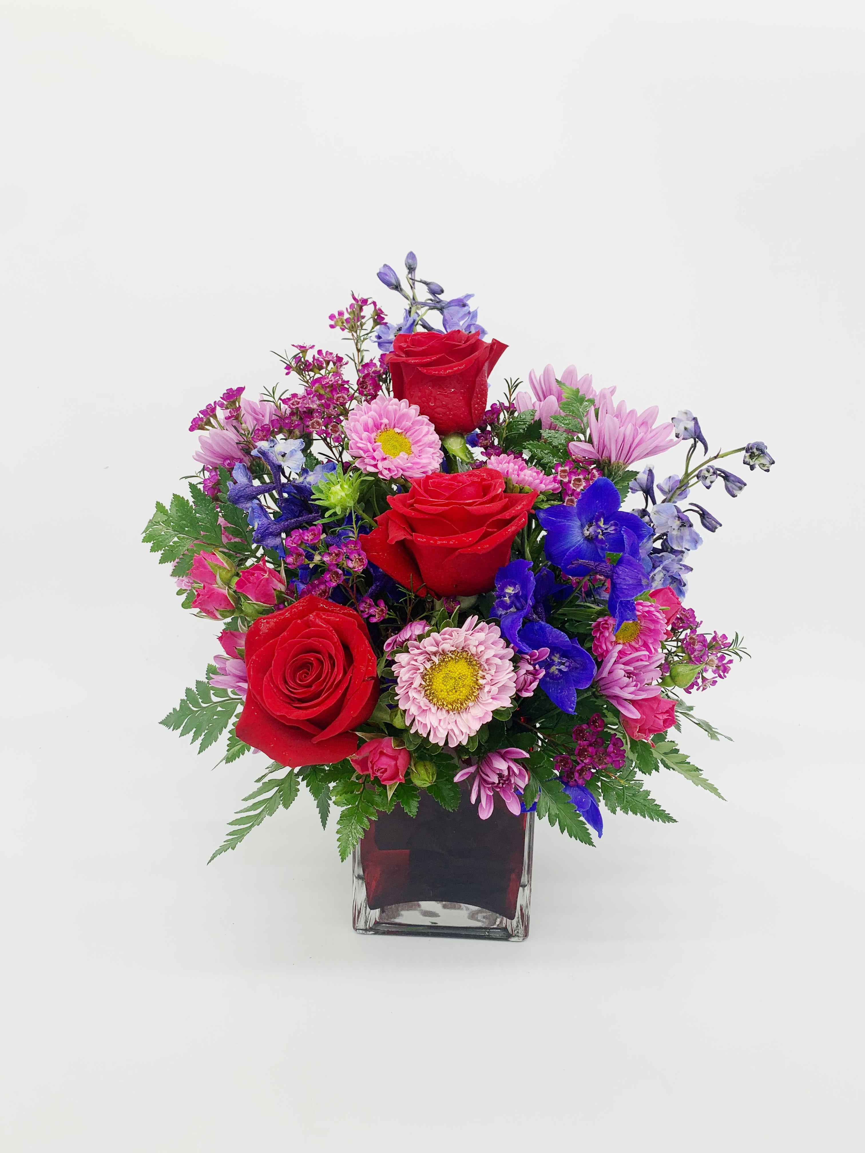 Vibrant mixed bouquet with red roses, pink daisies, and blue flowers in a red glass cube vase