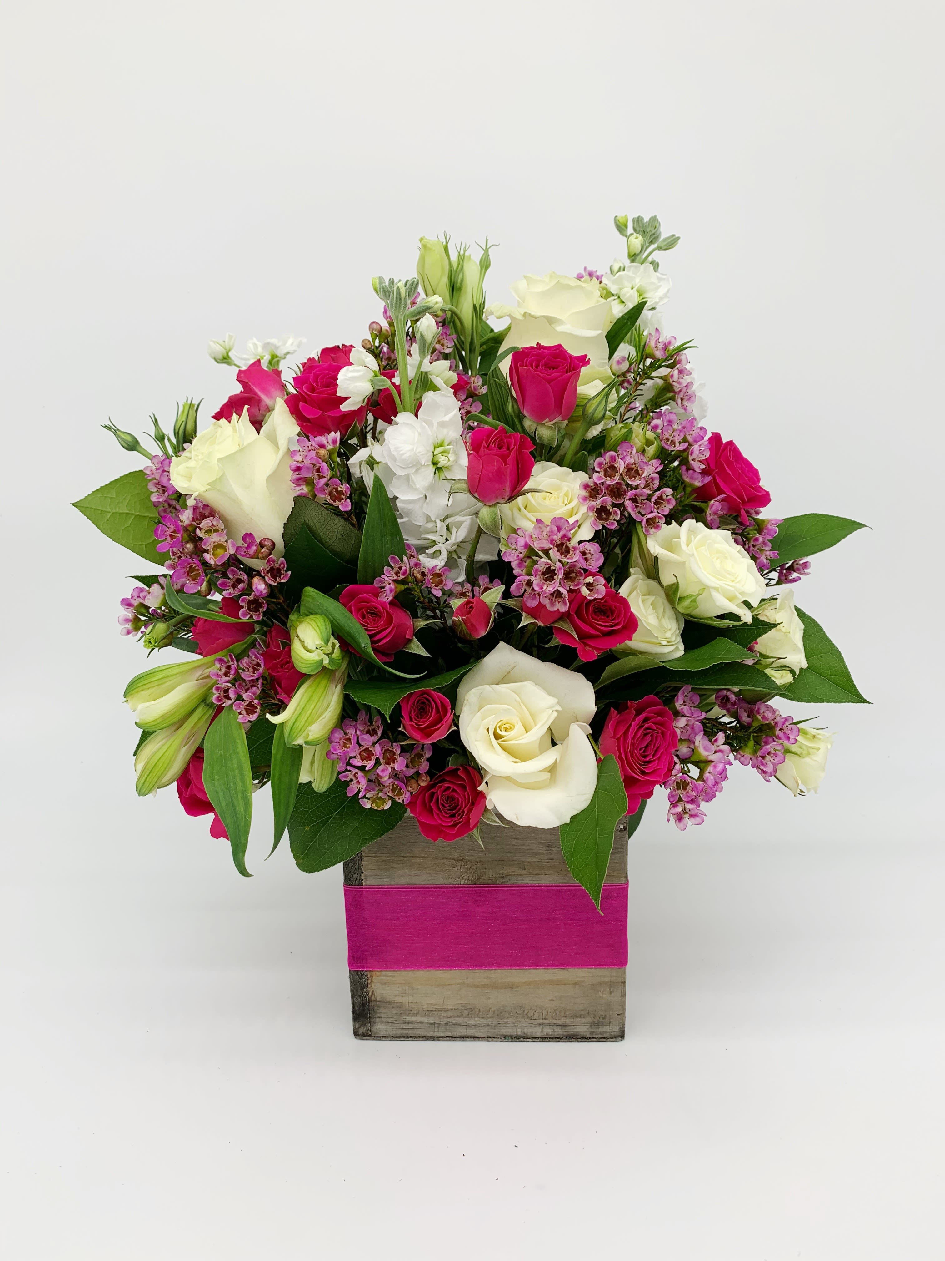 Compact arrangement of white and hot pink roses with small purple flowers in a wood cube vase wrapped with a pink band