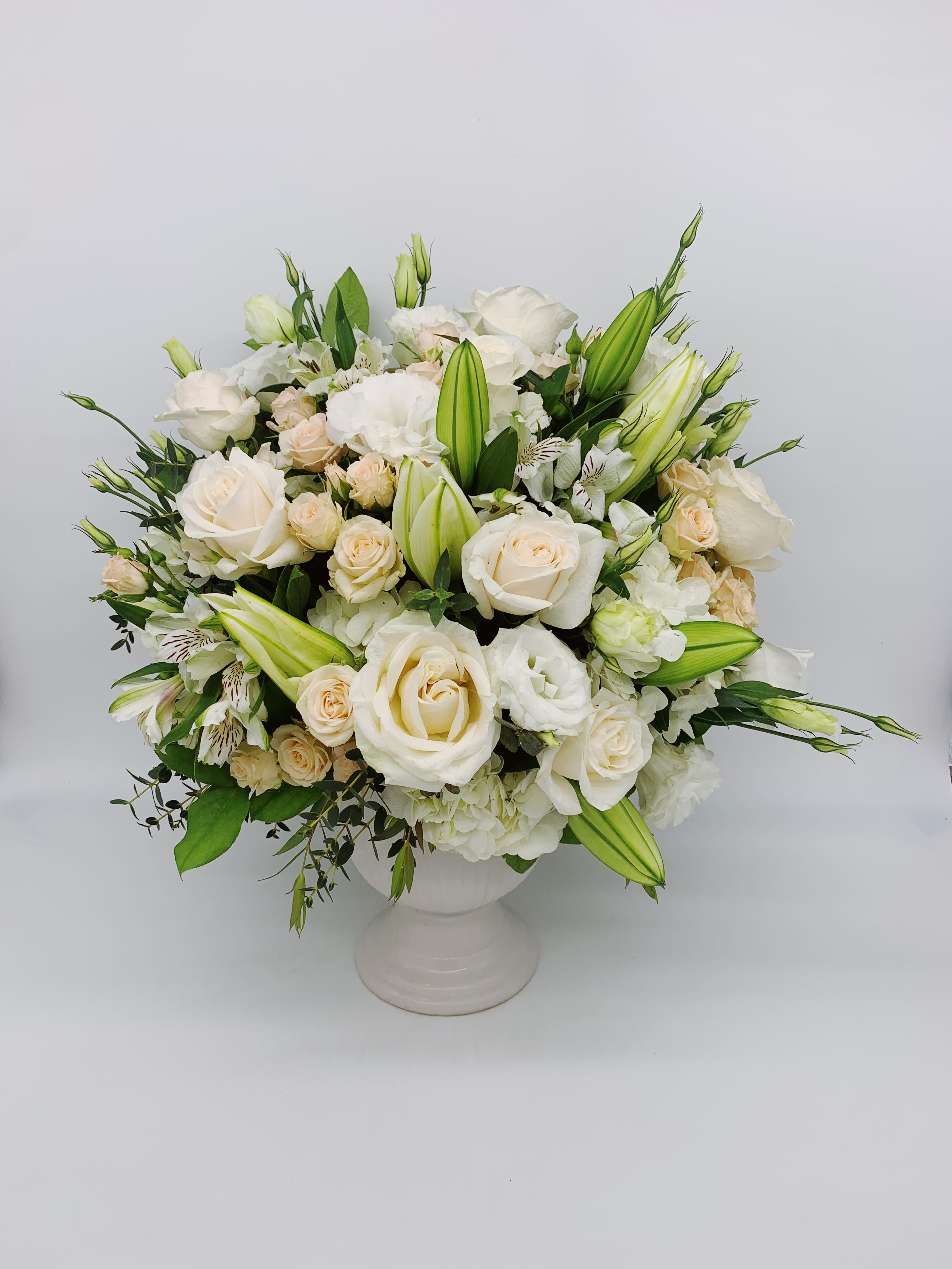 Round arrangement of white and cream roses, lilies, and mixed blooms in a white pedestal vase