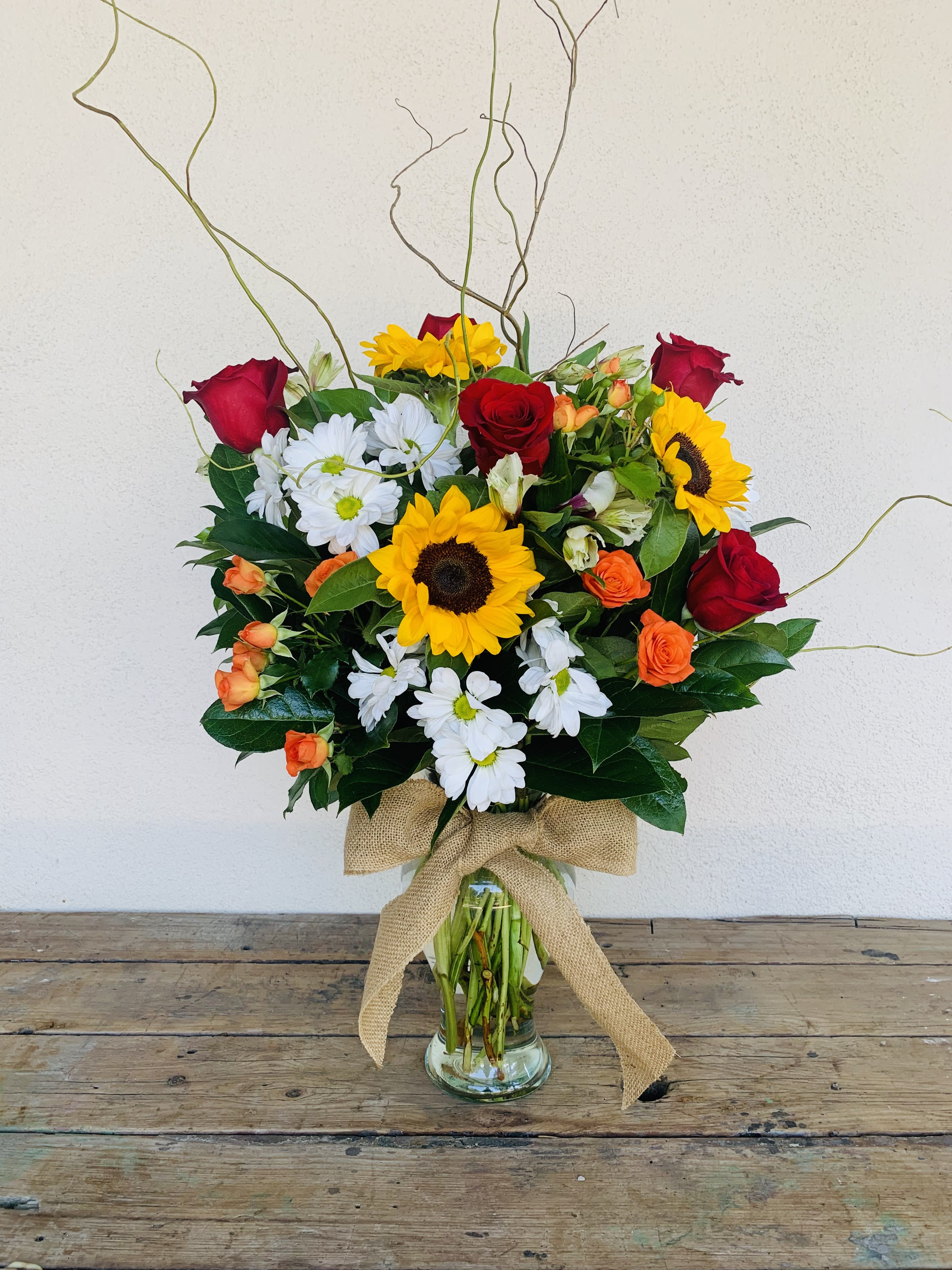 Tall mixed bouquet with sunflowers, red roses, white daisies, and orange blooms in a glass vase with burlap bow