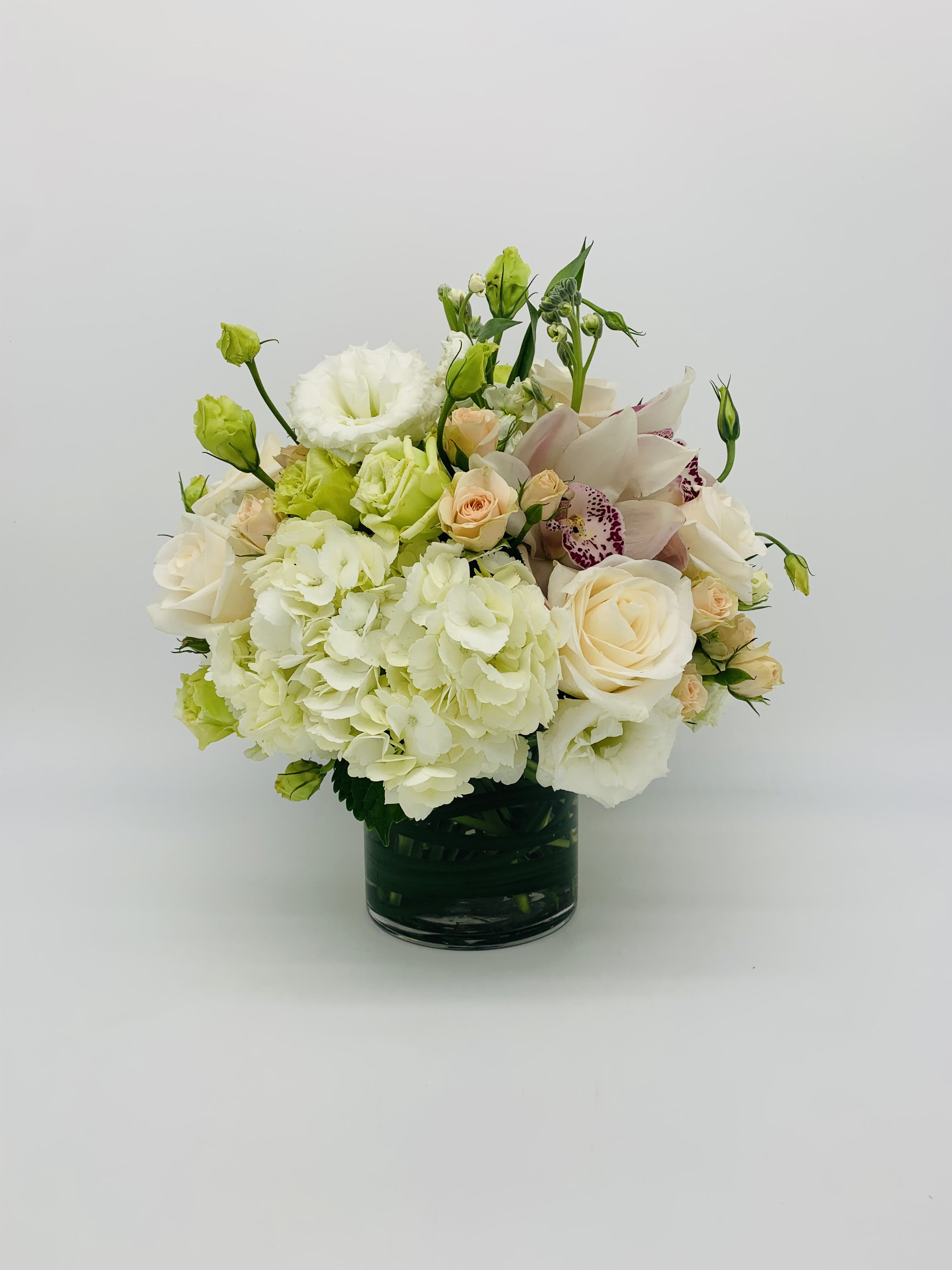 Round arrangement of white hydrangeas, cream roses, and orchids in a glass vase