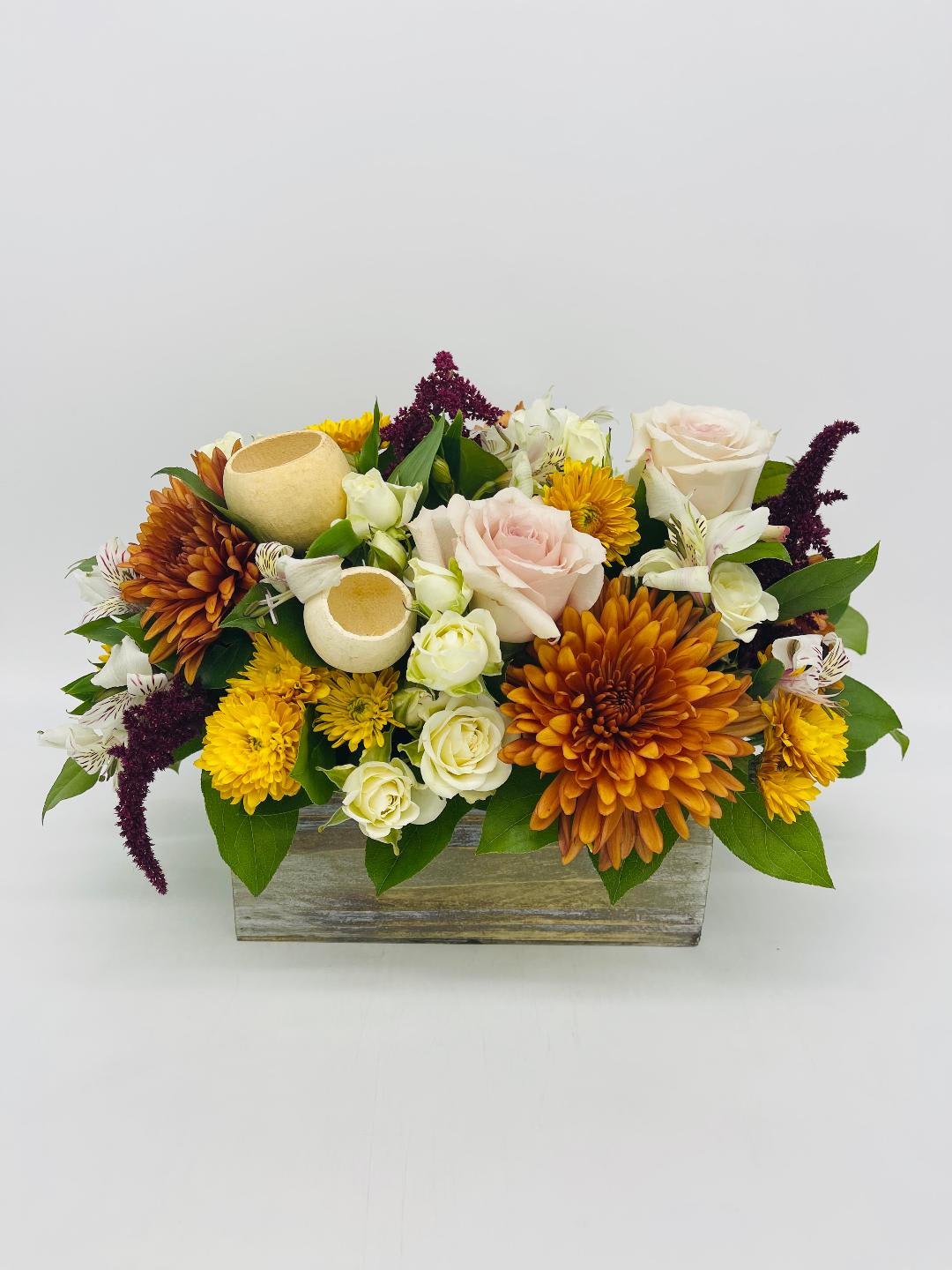 Low rectangular arrangement with orange and yellow mums, pale roses, and white blooms in a wooden box with decorative pods.