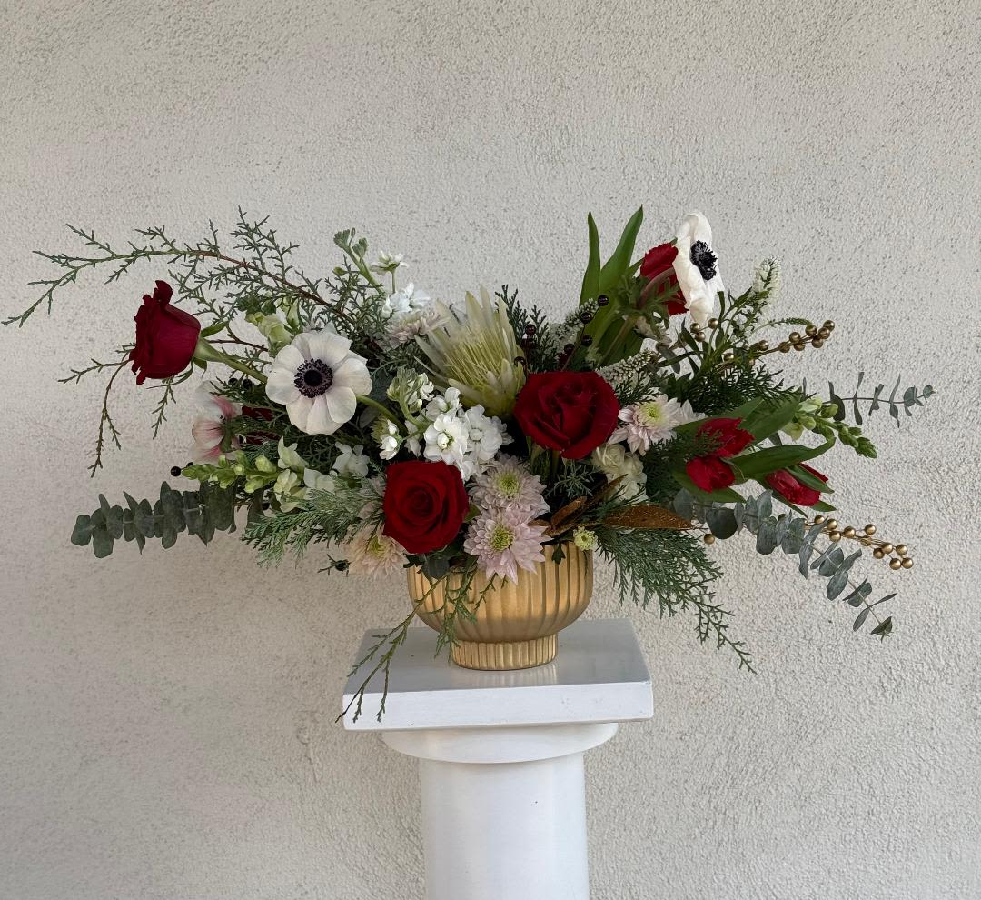 Asymmetrical arrangement of red roses and white flowers in a gold compote vase on a white pedestal.