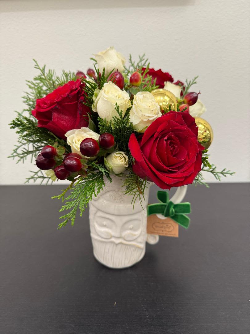 Compact arrangement of red and white roses with berries and evergreens in a white Santa mug.