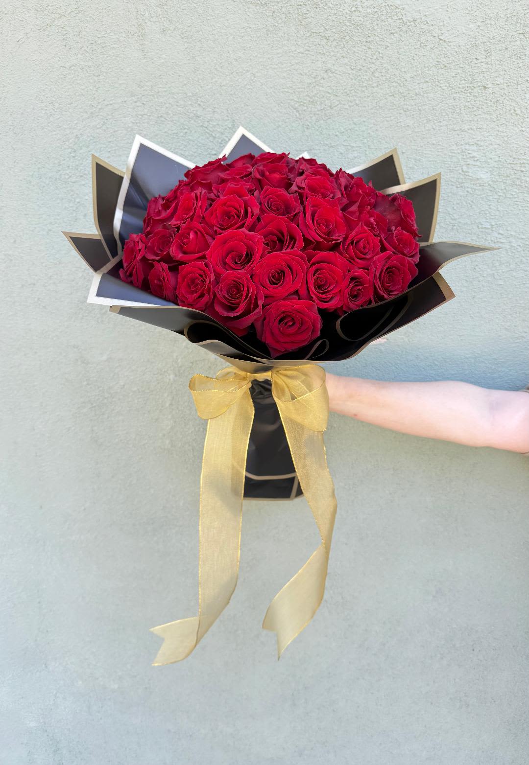 Arm holding a large bouquet of red roses wrapped in black paper with a gold bow.