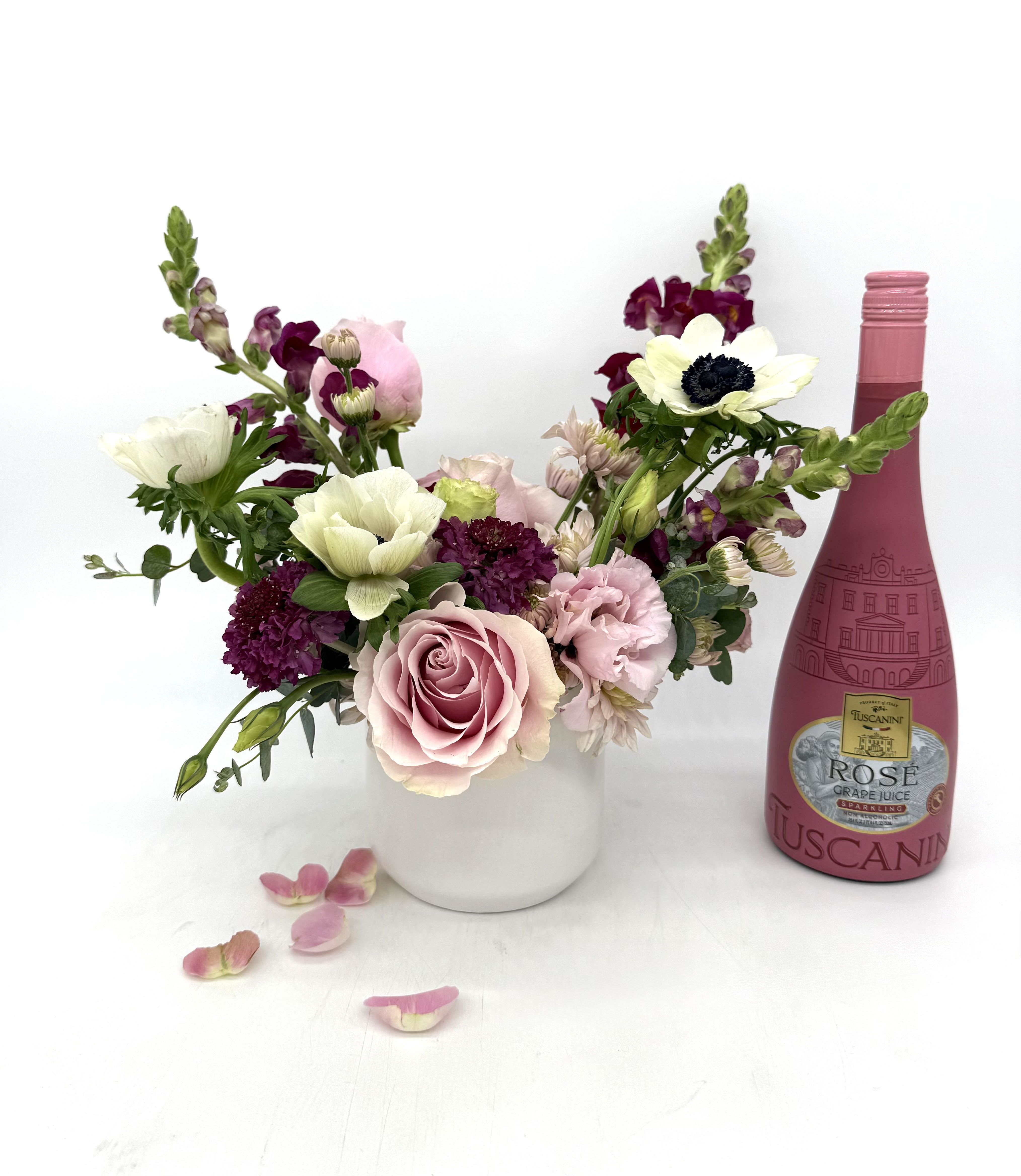 Mixed pink and white flower arrangement in white vase beside pink rosé bottle and scattered petals