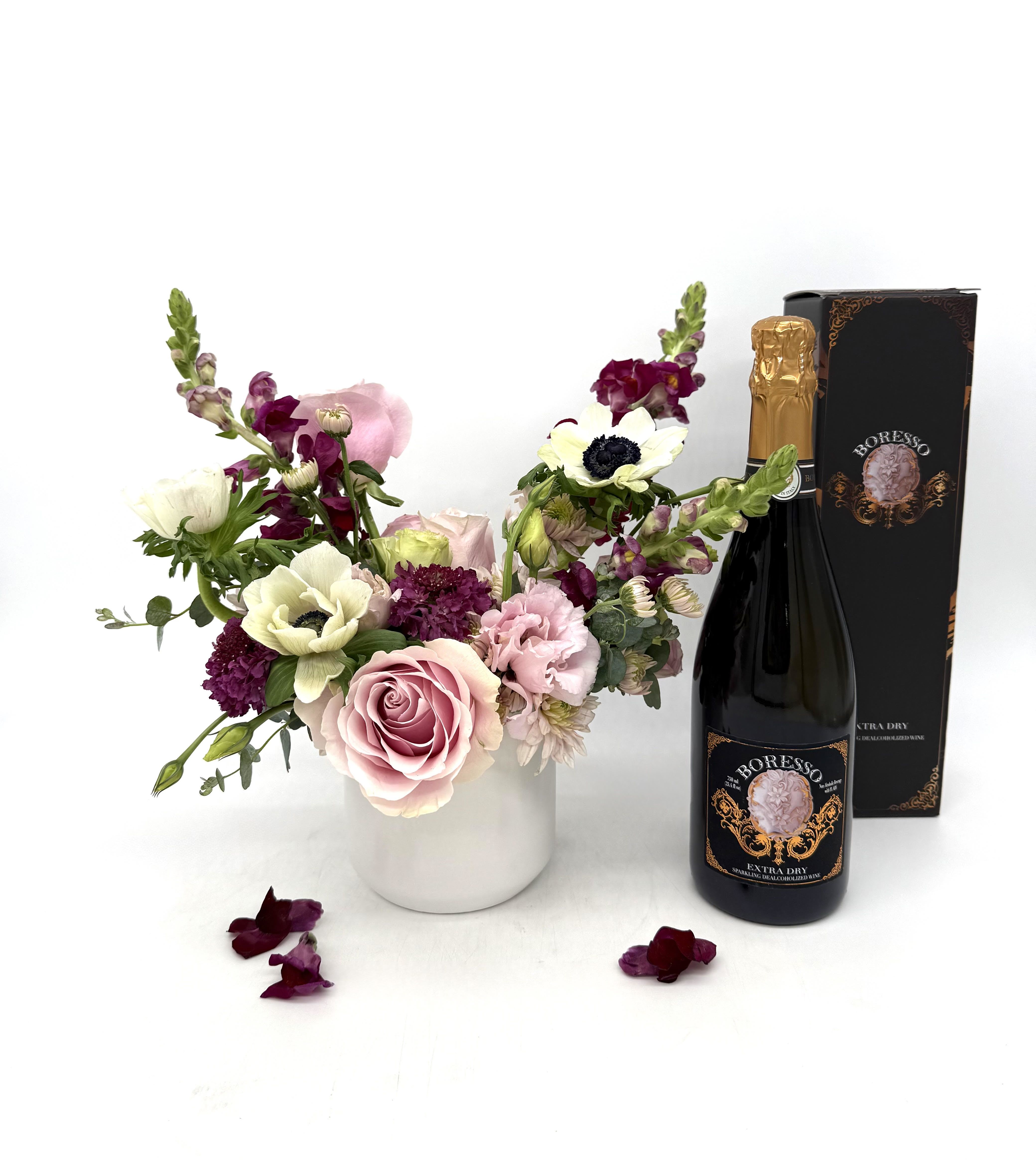 Pink and white floral arrangement in a white vase with a bottle of sparkling wine and gift box.