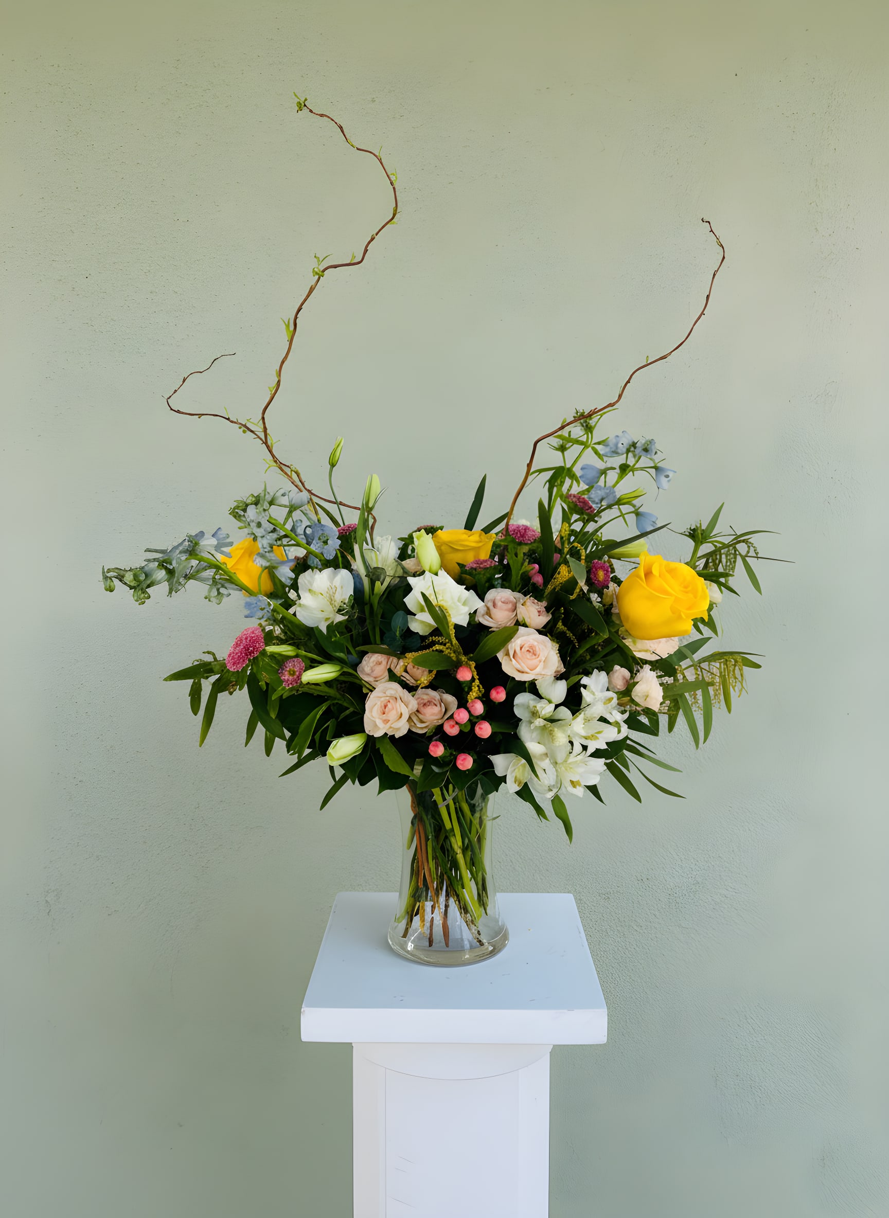 LF407 Sunshine Stems - Beautiful Spring vase arrangement, with yellow roses, blue delphinium, white lisianthus, pink asters, pops of pink spray roses and just the right amount of curly willow, sure to inspire all those spring feels. 