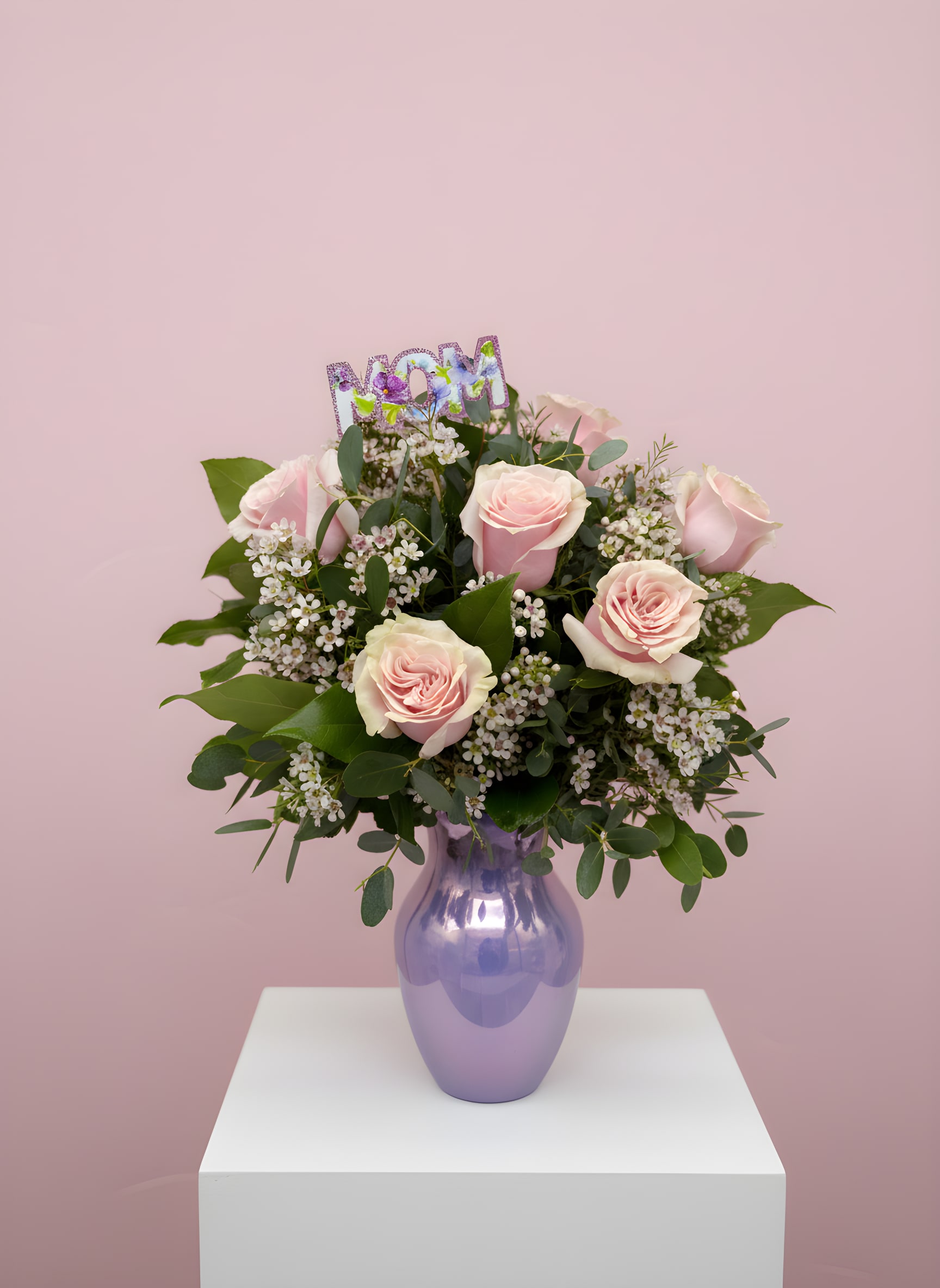LF412  Half Dozen for Mom - Celebrate Mom with this elegant arrangement featuring soft blush roses, white wax flowers, and lush greenery, all beautifully displayed in a radiant lavender vase.  The shimmering "MOM" topper adds a heartfelt touch.  It's the perfect gift to brighten her day and express your love this Mother's Day 