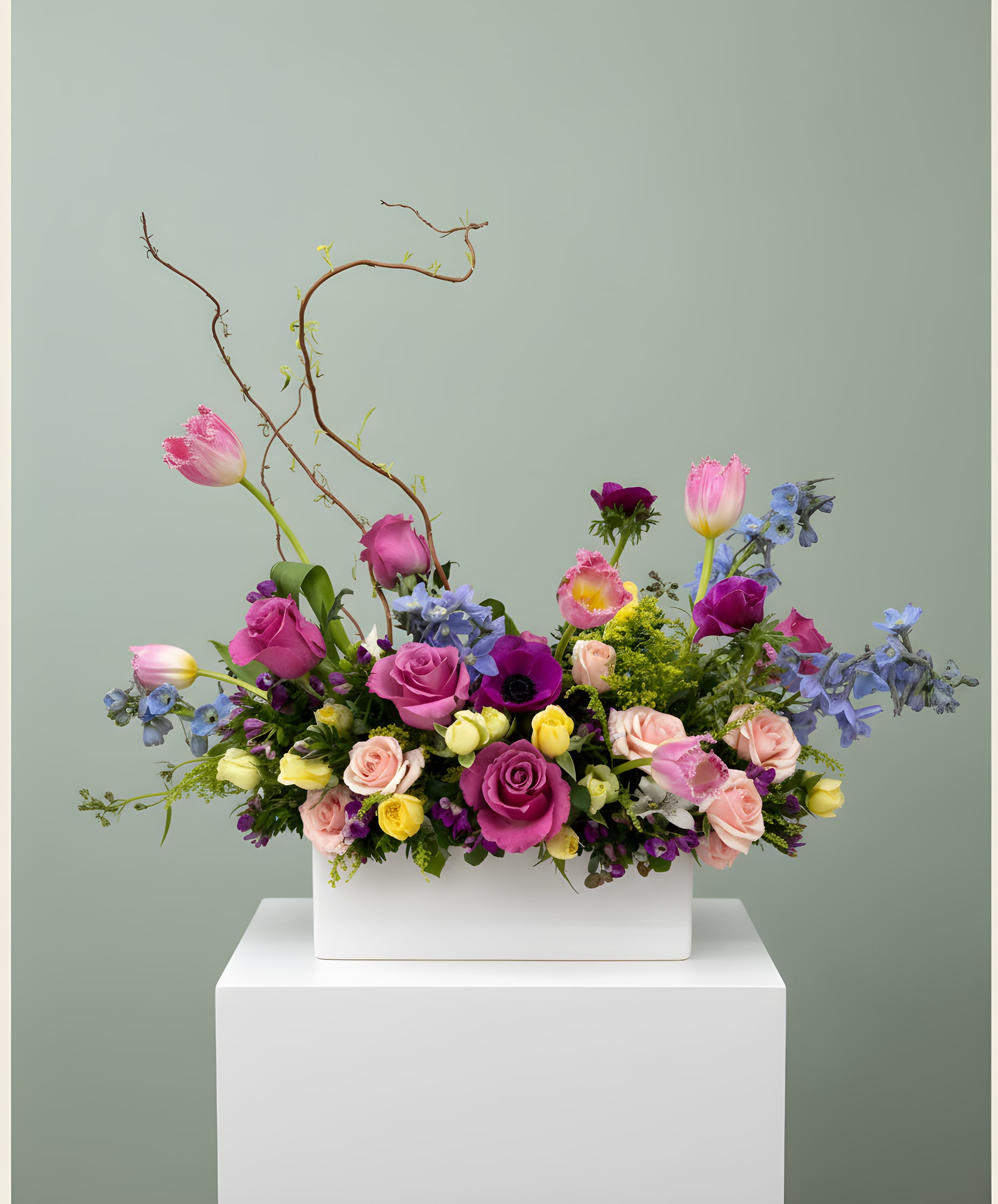 LF 395  Queen Bee - This beautiful bouquet will surely delight the Queen in your life.  Lovely spring blooms which include roses, delphinium, tulips, anemone, curly willow and an array of other spring mixes. All this beauty in a white ceramic container is perfect for a Queen.   Orientation- All Around Approx Dim.  16" W x 12" H x 8" D