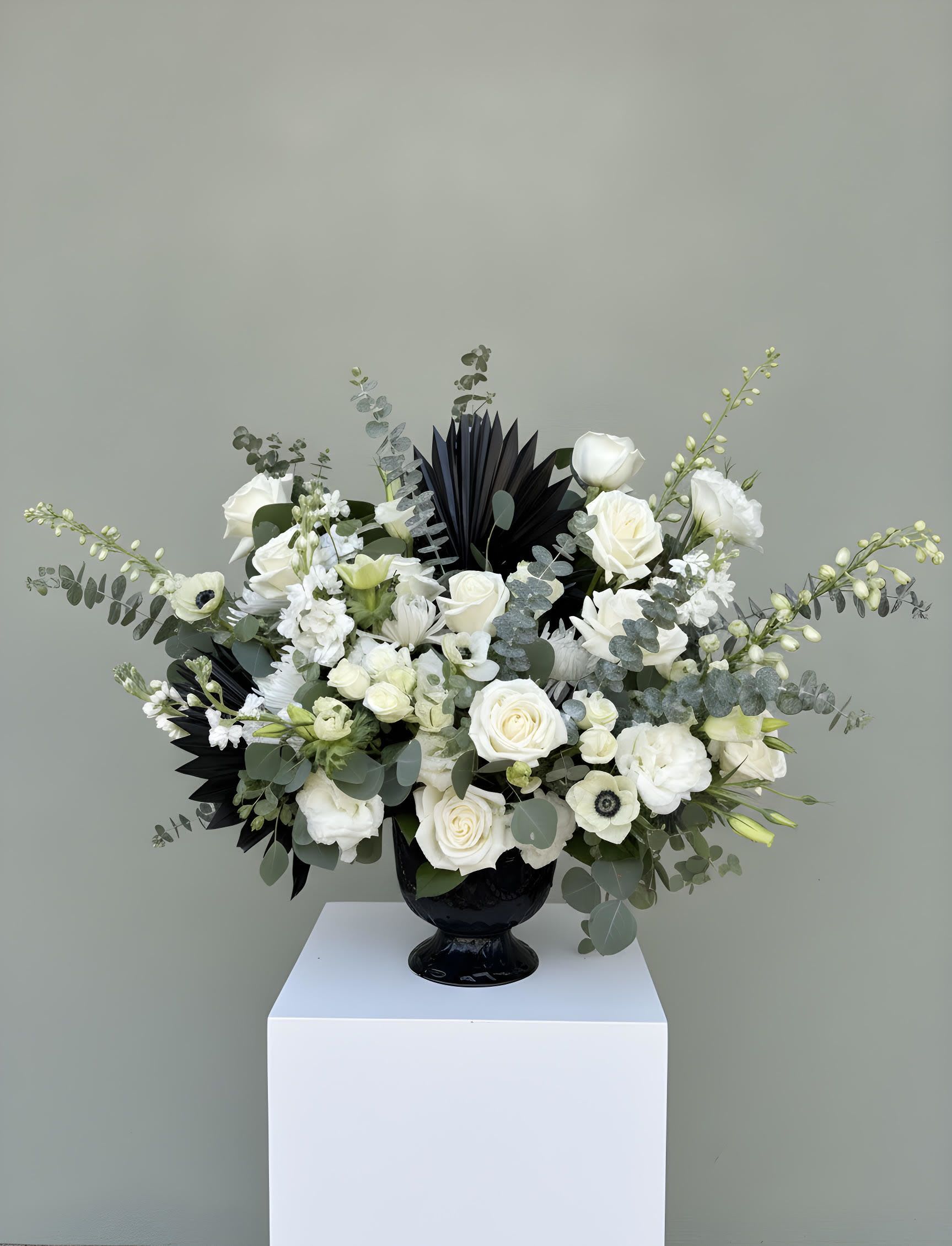 LF422  Midnight Grace Alter Piece - A striking and elegant funeral altar arrangement featuring lush white roses, lisianthus, anemones, and delicate seasonal blooms accented with eucalyptus and dramatic black palm foliage.  Designed in a sophisticate black vessel, this contemporary tribute offers a beautiful expression of love, remembrance, and honor to a memorial service. 