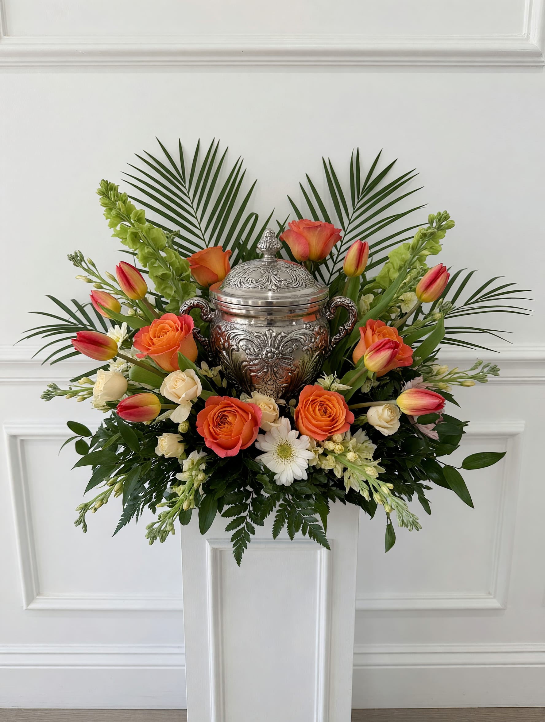 LF423 Sunset Remembrance Urn Tribute - A warm and uplifting urn arrangement featuring vibrant orange roses, tulips, creamy white blooms with lush greenery and palms surrounding a cherished urn.  Designed to celebrate a life well lived, this elegant tribute brings comfort, beauty and a sense of peaceful remembrance to any memorial service