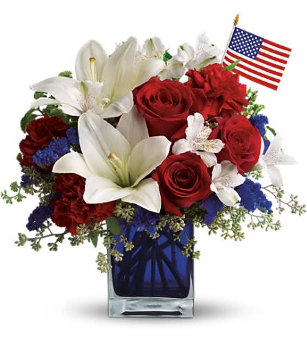 Red, white, and blue flower arrangement with roses and lilies in a blue vase with a small American flag.