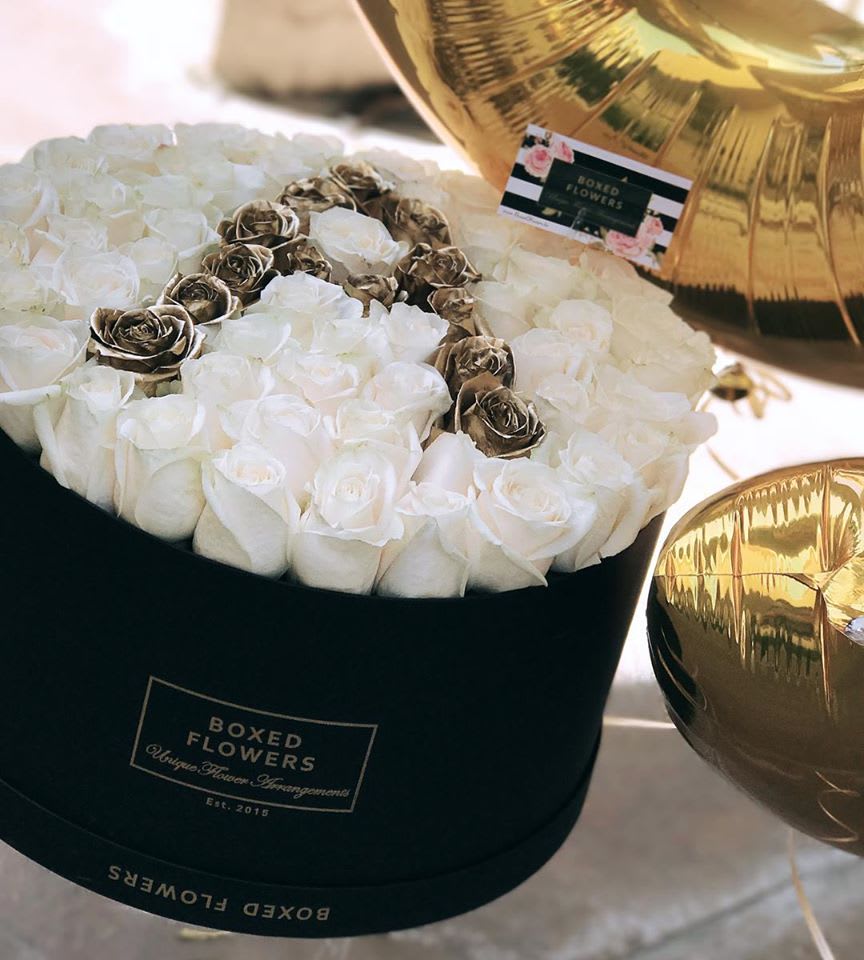 White roses arranged in a black round box with gold balloons nearby.