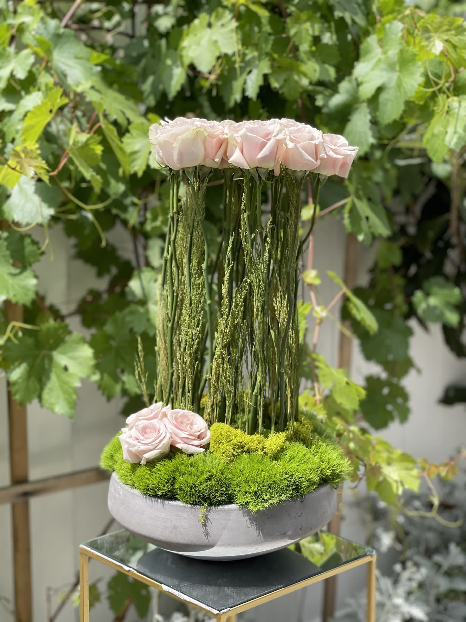 Pink roses arranged in a tall moss-filled bowl with hanging greenery