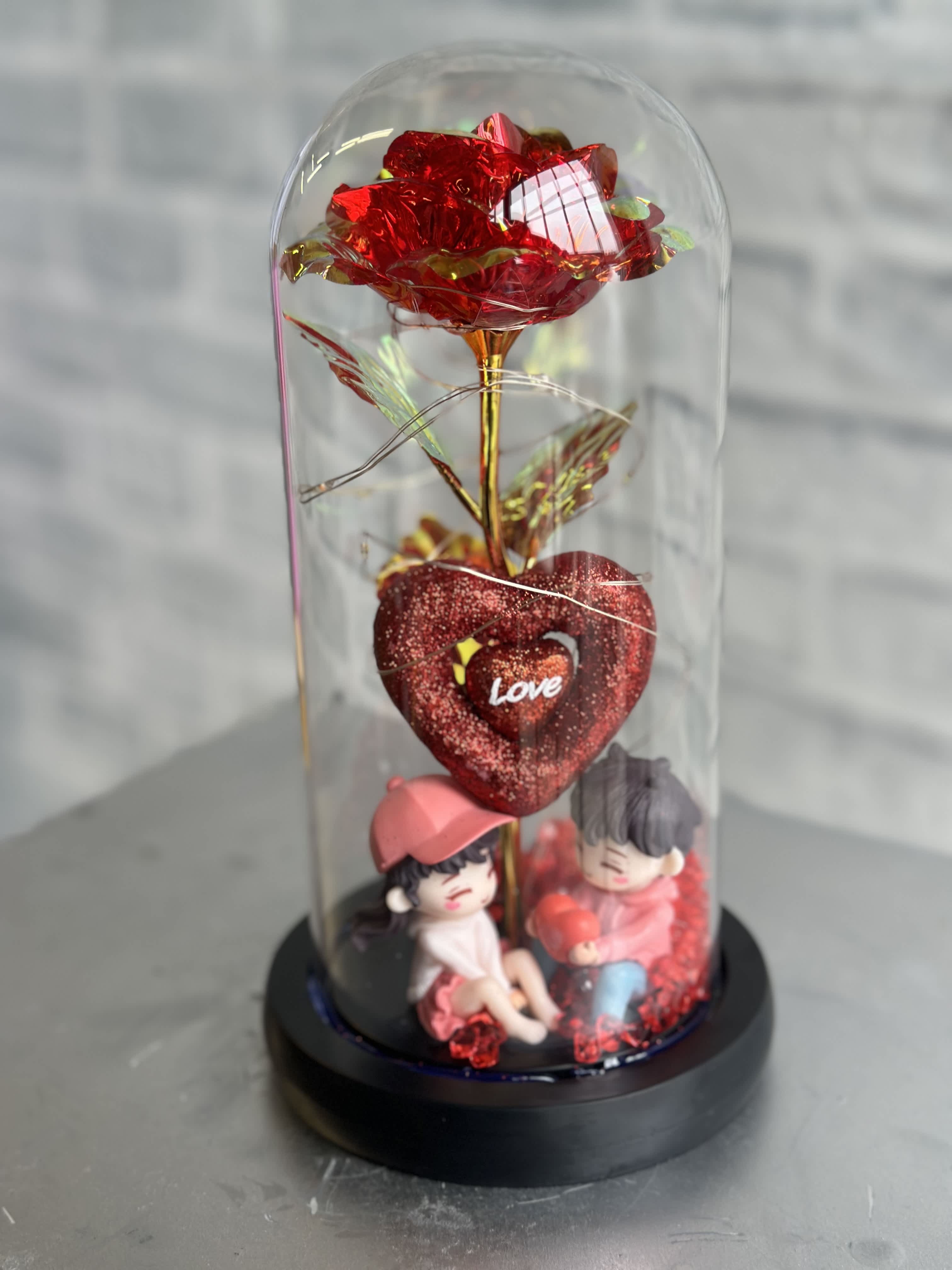 Red decorative rose under a clear dome with two small figurines at the base