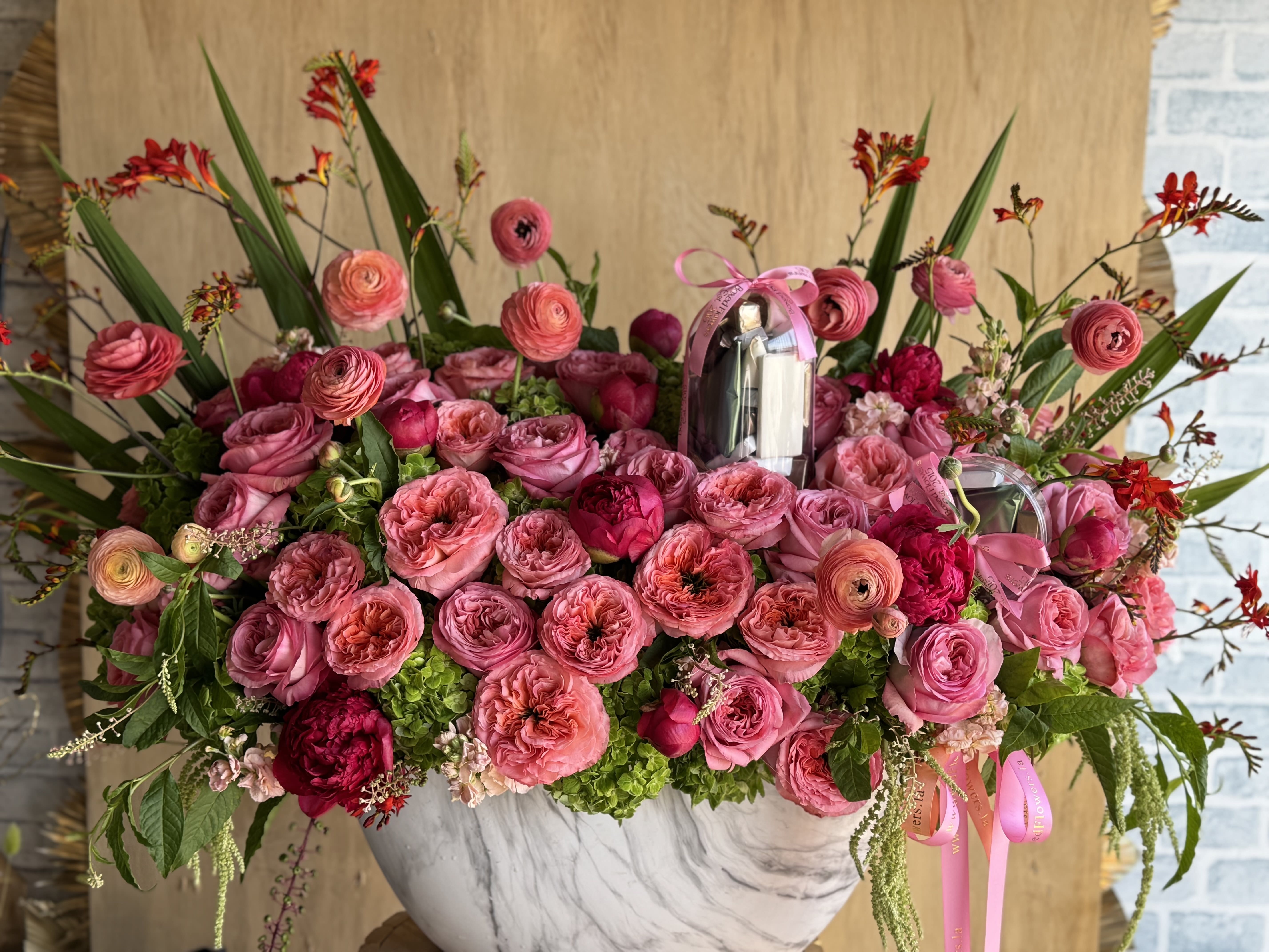 Pink Reverie - A dazzling dream in every shade of pink, Pink Reverie is an opulent floral arrangement bursting with lush blooms and thoughtful detail. Featuring a luxurious blend of garden roses, ranunculus, peonies, and bright green hydrangea, this design is accented with bold red crocosmia and tropical greenery for height and drama. Nestled among the blooms is a delightful surprise—1.5 pounds of premium assorted chocolates, elegantly wrapped for gifting. Presented in a marble-textured ceramic vessel, this arrangement is perfect for romantic occasions, heartfelt celebrations, or any moment worth indulging.