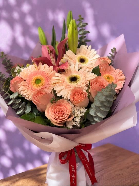 THANK YOU - A graceful hand-tied bouquet of peach gerberas, salmon roses, pink lilies, and eucalyptus, wrapped in blush paper and finished with a red ribbon. A sophisticated gift for any occasion.
