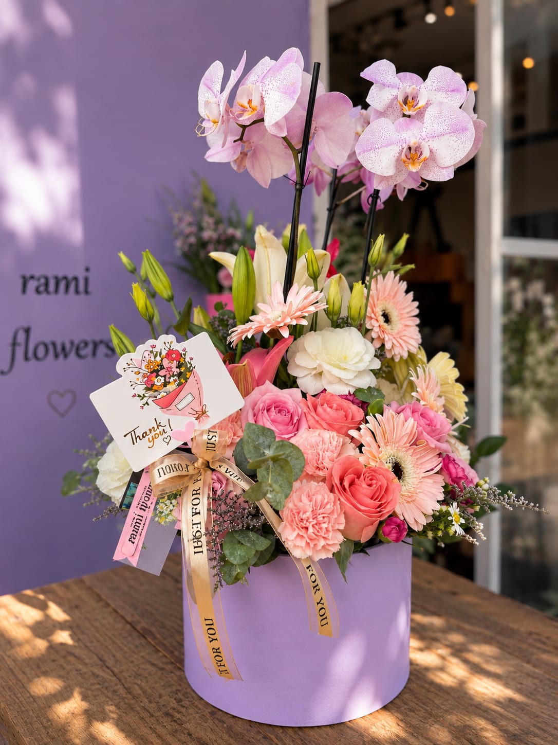 NEW BEGINNINGS - A stunning tall arrangement in a lavender cylinder box featuring elegant phalaenopsis orchids rising above a lush mix of peach roses, pink gerberas, carnations, white eustoma, and lily buds. The ultimate premium gift.