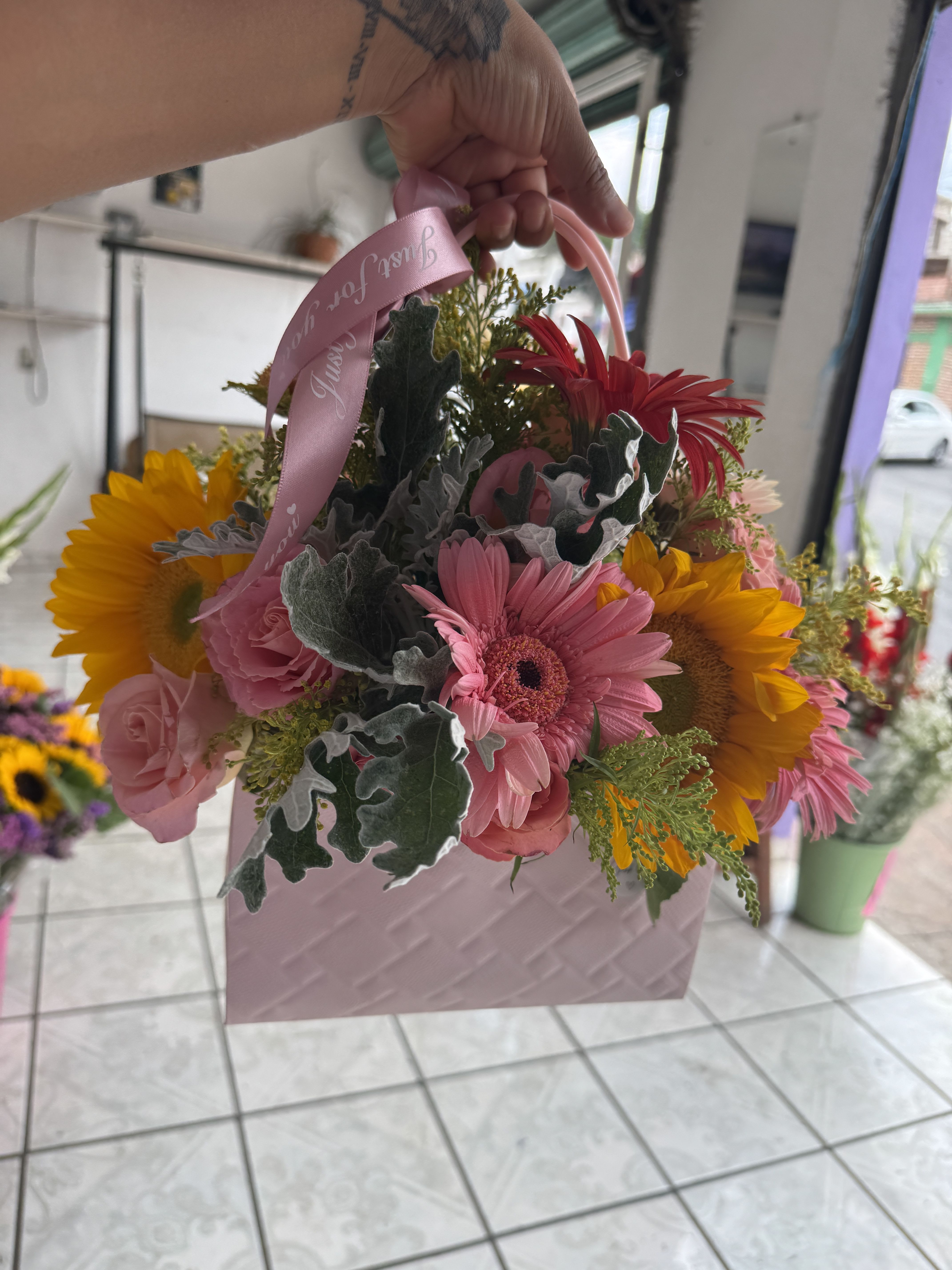 HAPPY BIRTHDAY - Flowers meet fashion in this utterly charming arrangement — a lush mix of golden sunflower, pink and red gerbera daisies, soft pink roses, dusty miller, and yellow solidago, all arranged in a chic quilted pink handbag-style keepsake box, finished with a personalized satin ribbon handle. The bag is theirs to keep and reuse long after the last bloom fades — a gift within a gift. Fun, feminine, and completely one-of-a-kind, this is the perfect surprise for Mom, a birthday girl, a teacher, or anyone who loves flowers with a side of style. 