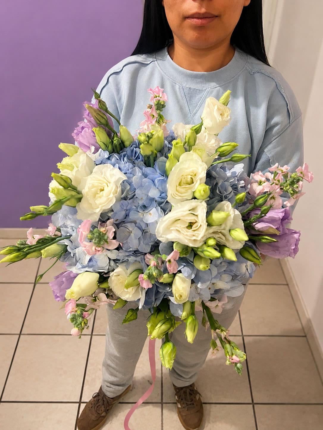 SOMETHING BLUE  - Soft, serene, and utterly romantic — this oversized garden bouquet is a dream in blue and white. Pillowy blue hydrangeas form a lush, cloud-like base, crowned with creamy white garden roses in full bloom and accented by lavender lisianthus and delicate pink stock flowers. Every stem tells a story of elegance and effortless style. Generously sized and hand-tied with a whisper-pink trailing ribbon, this bouquet makes a breathtaking statement — whether carried down the aisle or gifted to someone who deserves the very best.