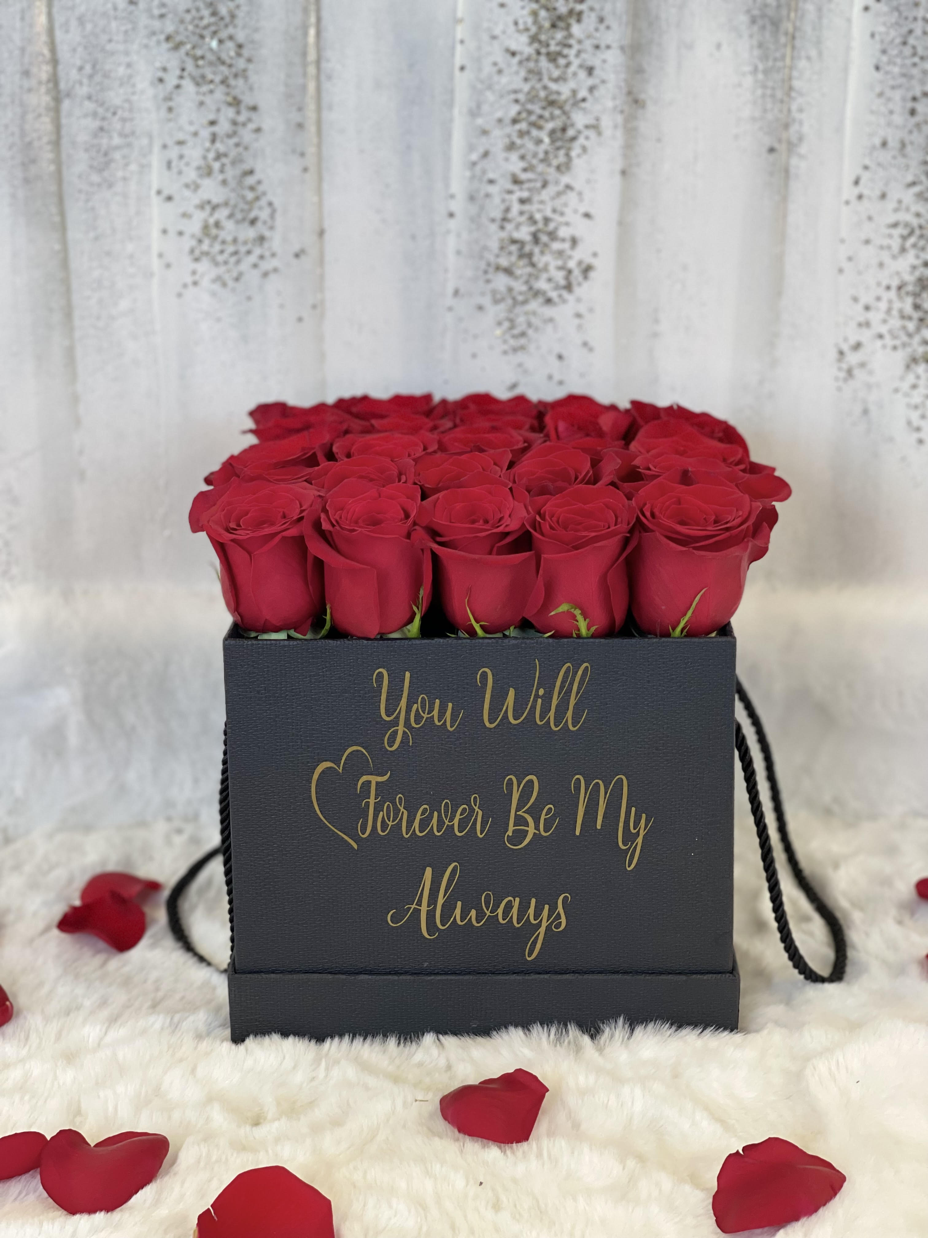 Square black box of red roses with gold script text, surrounded by scattered red rose petals
