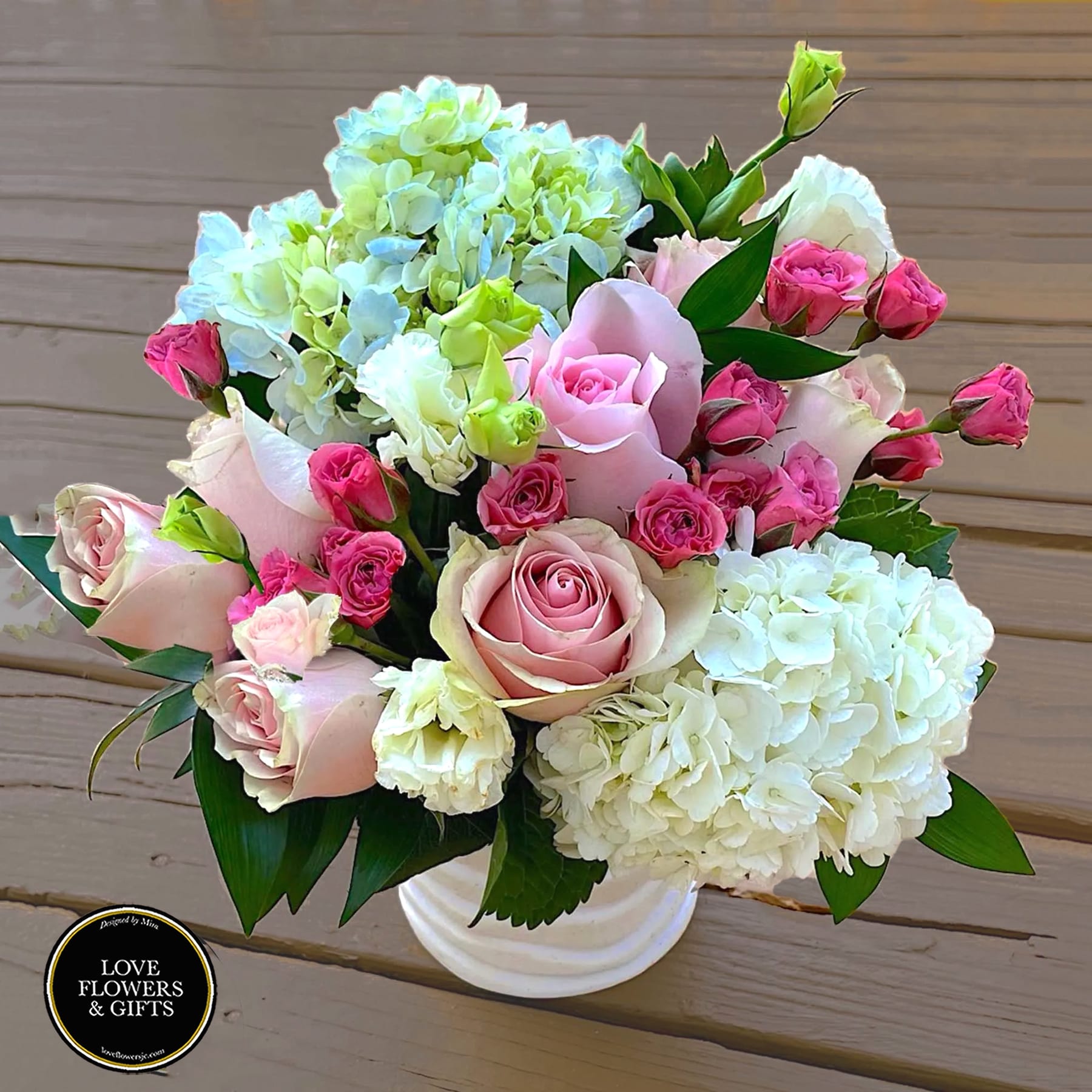 Compact arrangement of pink roses and spray roses with white and green hydrangeas in a white vase