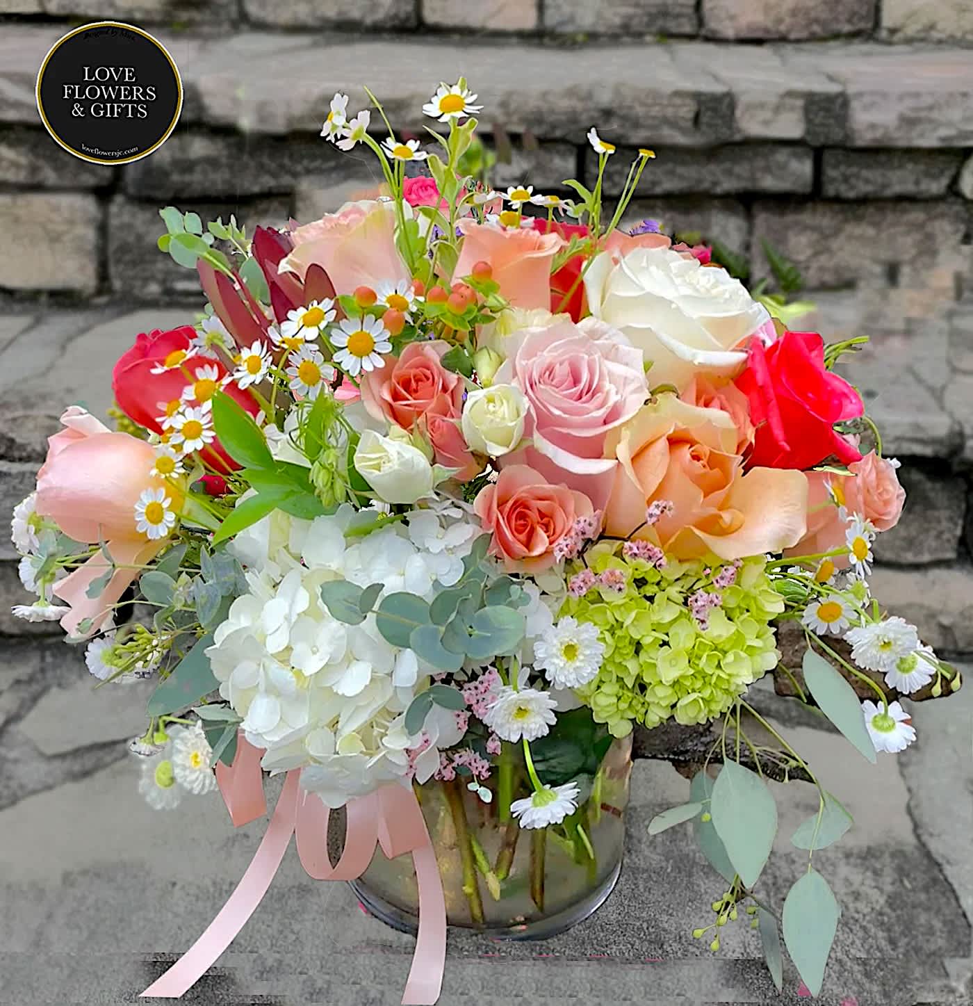 Low glass vase of mixed roses, hydrangeas, and daisies in soft pink, peach, white, and green tones