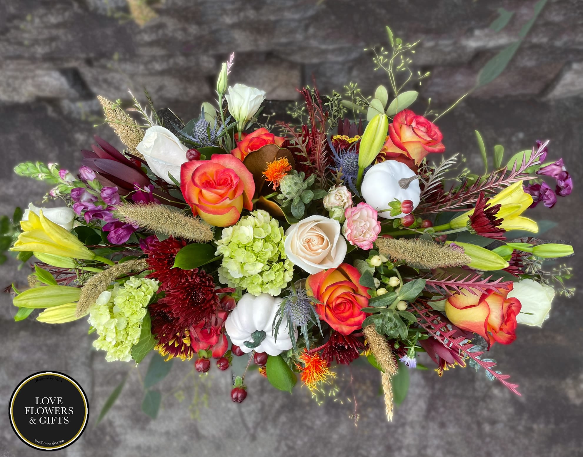 Elongated fall flower arrangement with orange roses, yellow lilies, green hydrangeas, and white pumpkins.