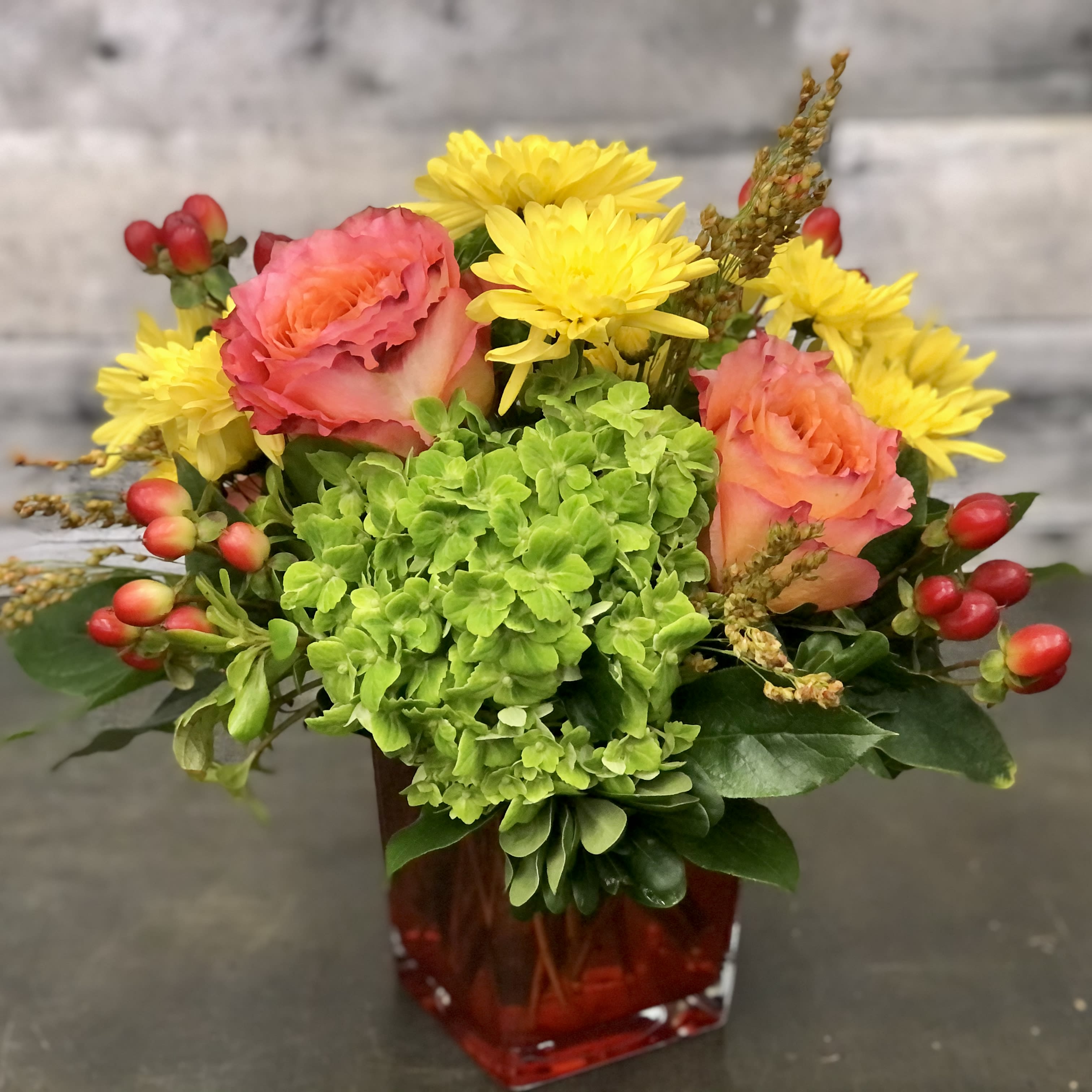 Alicia  - Vibrant roses and mums in a hydrangea bed with hypericum and sorghum accents all set in an orange cube. Perfect for fall or any season!
