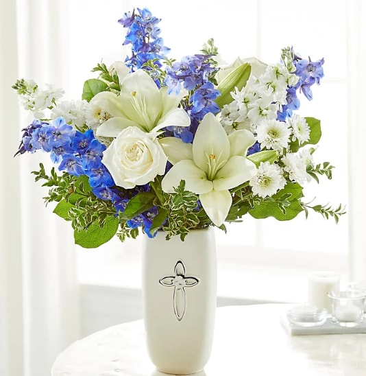 White lilies, roses, and blue flowers in a white vase with a silver cross design