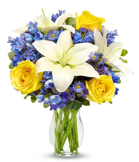 Clear vase of white lilies, yellow roses, and blue flowers in a rounded mixed bouquet