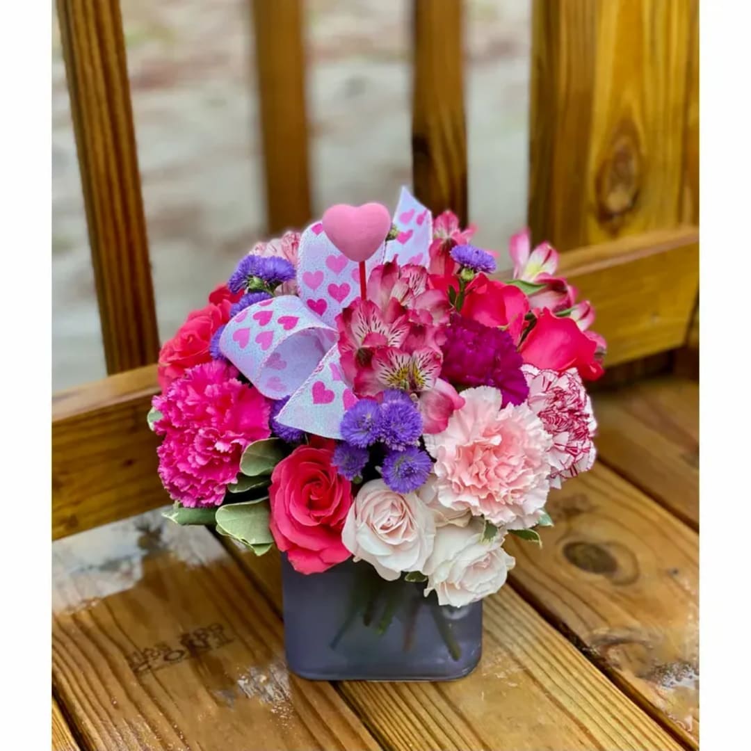 Pink and purple mixed flower arrangement in a square vase with heart decorations