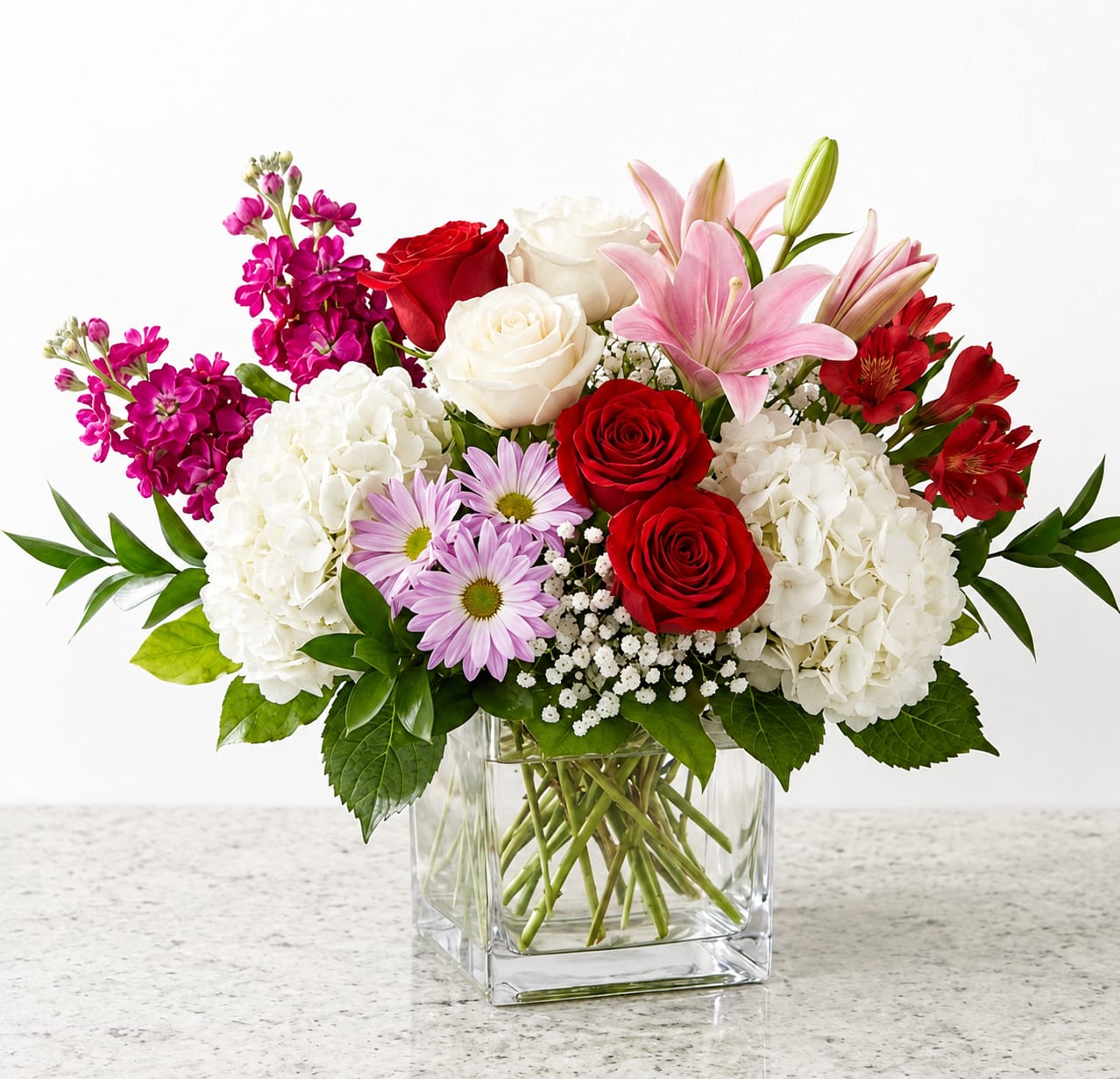 Cutesy Cube - A lush mix of red and white roses with pink lilies takes center stage in this vibrant birthday flower arrangement, designed to make the person you're thinking of feel cherished. The combination reads romantic enough for an anniversary or love-and-romance moment, yet fresh and cheerful for a just-because surprise or a heartfelt thank-you.  Creamy white hydrangea clusters anchor the sides while purple daisy-like blooms and red alstroemeria weave color and texture through the middle; stock adds soft vertical accents for fragrance and fullness. Arranged in a clear square glass vase, the composition is balanced with taller lilies giving graceful height and rounded blooms creating a generous, gift-worthy silhouette.  Perfect for celebrating milestones or brightening an ordinary day, this arrangement arrives ready to display on a table or entryway. For best lasting beauty, keep the vase filled with fresh water and trim stems slightly when refreshing the arrangement; available in a single well-filled size ideal for gifting.