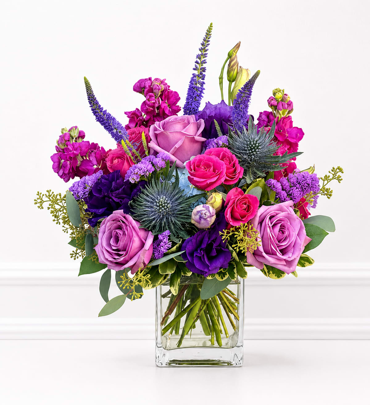 Perfect Royal - A lush mix of purple, pink, white and soft green florals creates a richly layered bouquet that feels both joyful and reverent - perfect as a birthday gift or a "just because" surprise that says you care. This flower arrangement for birthdays, anniversaries, or thinking-of-you moments combines rounded roses and spray roses with airy lisianthus, ruffled carnations, and tall stock stems to give height and movement; the color story leans on romantic pinks and saturated purples softened by ivory blooms and fresh greenery.  Arranged in a clear glass vase, the bouquet balances full, garden-style clusters with upright spiky accents for texture and dimension.