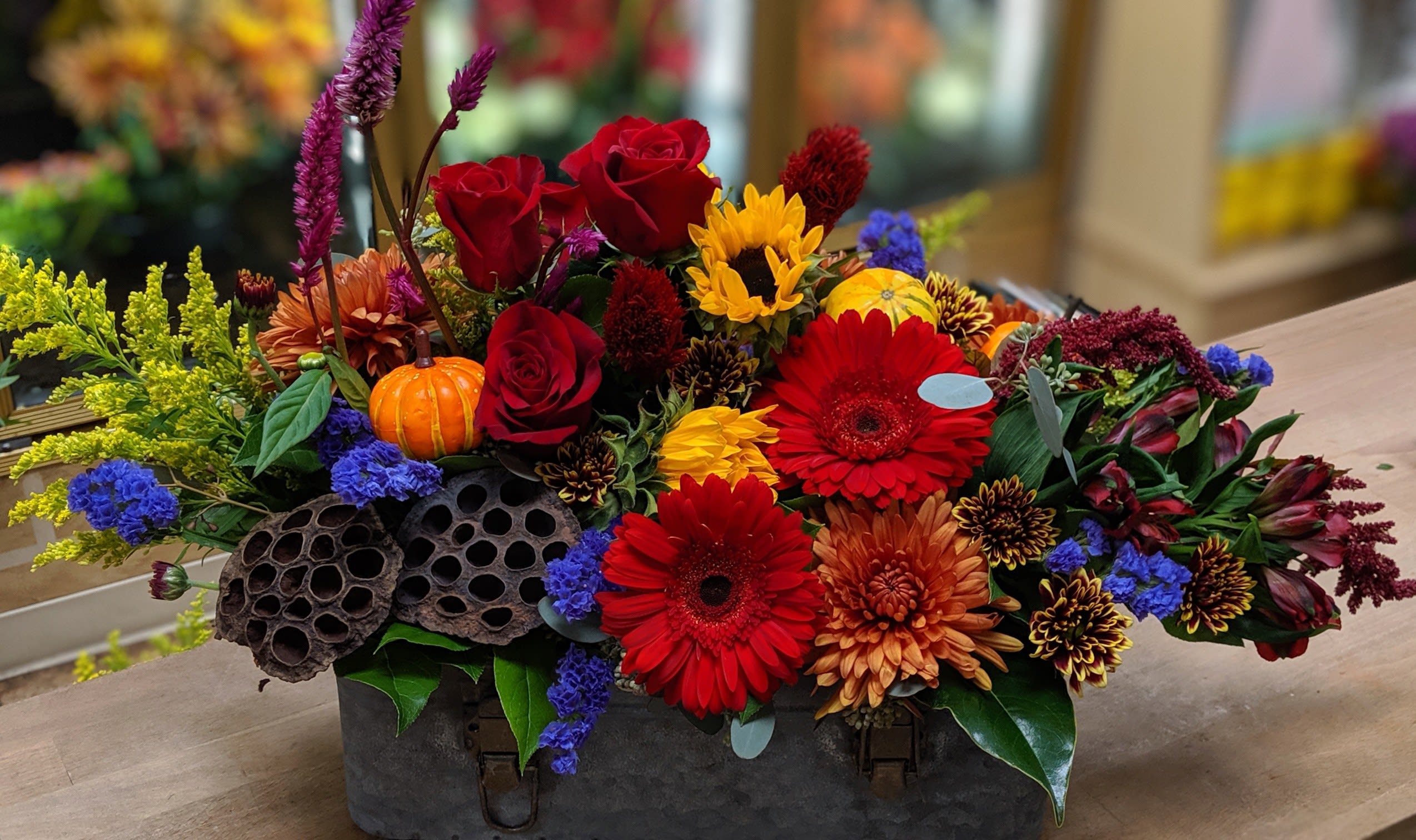 Colorful floral arrangement with roses, gerbera daisies, sunflowers, and a small pumpkin in a metal container