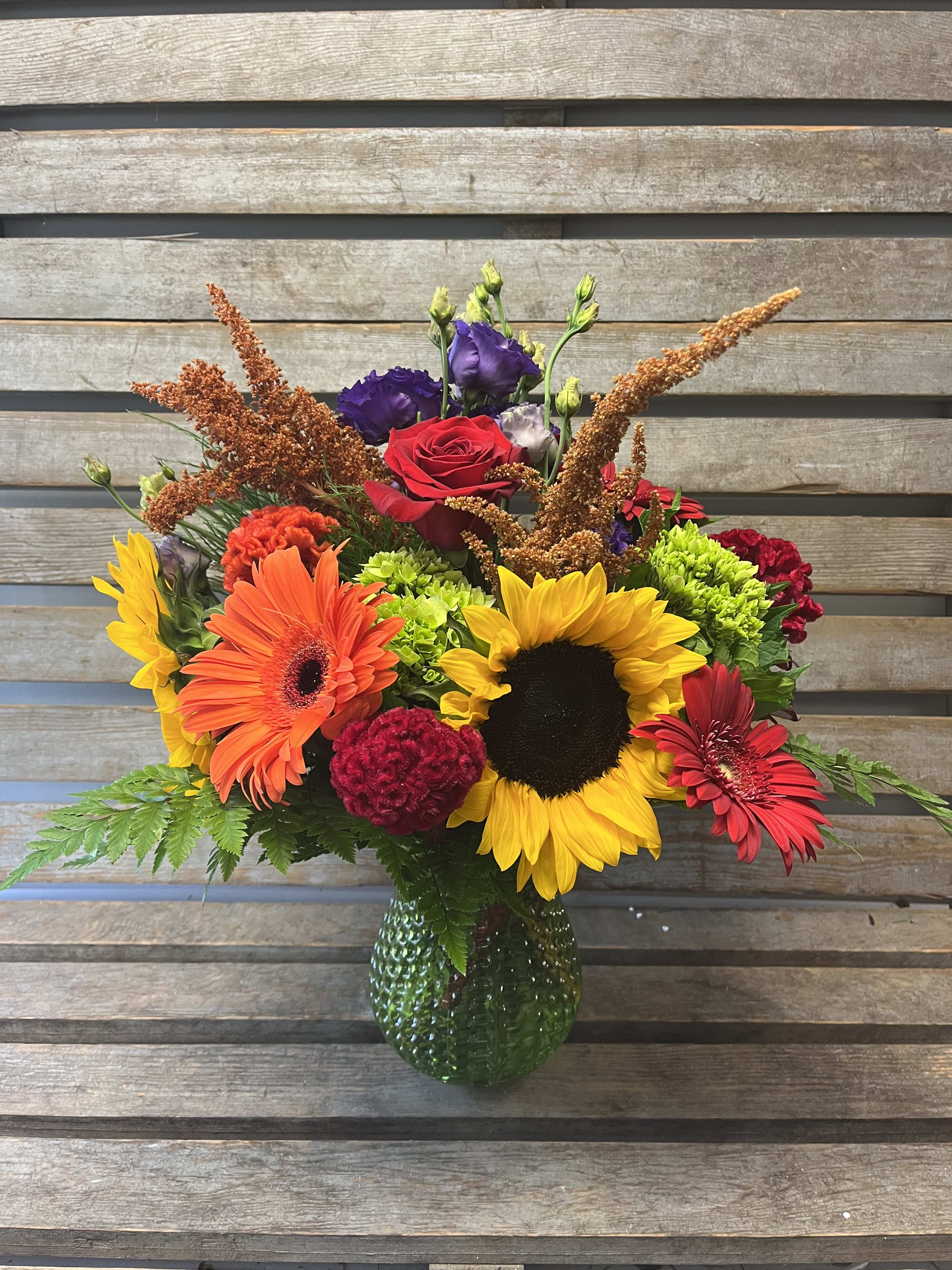 Colorful mixed bouquet with sunflowers, gerbera daisies, and roses in a green glass vase