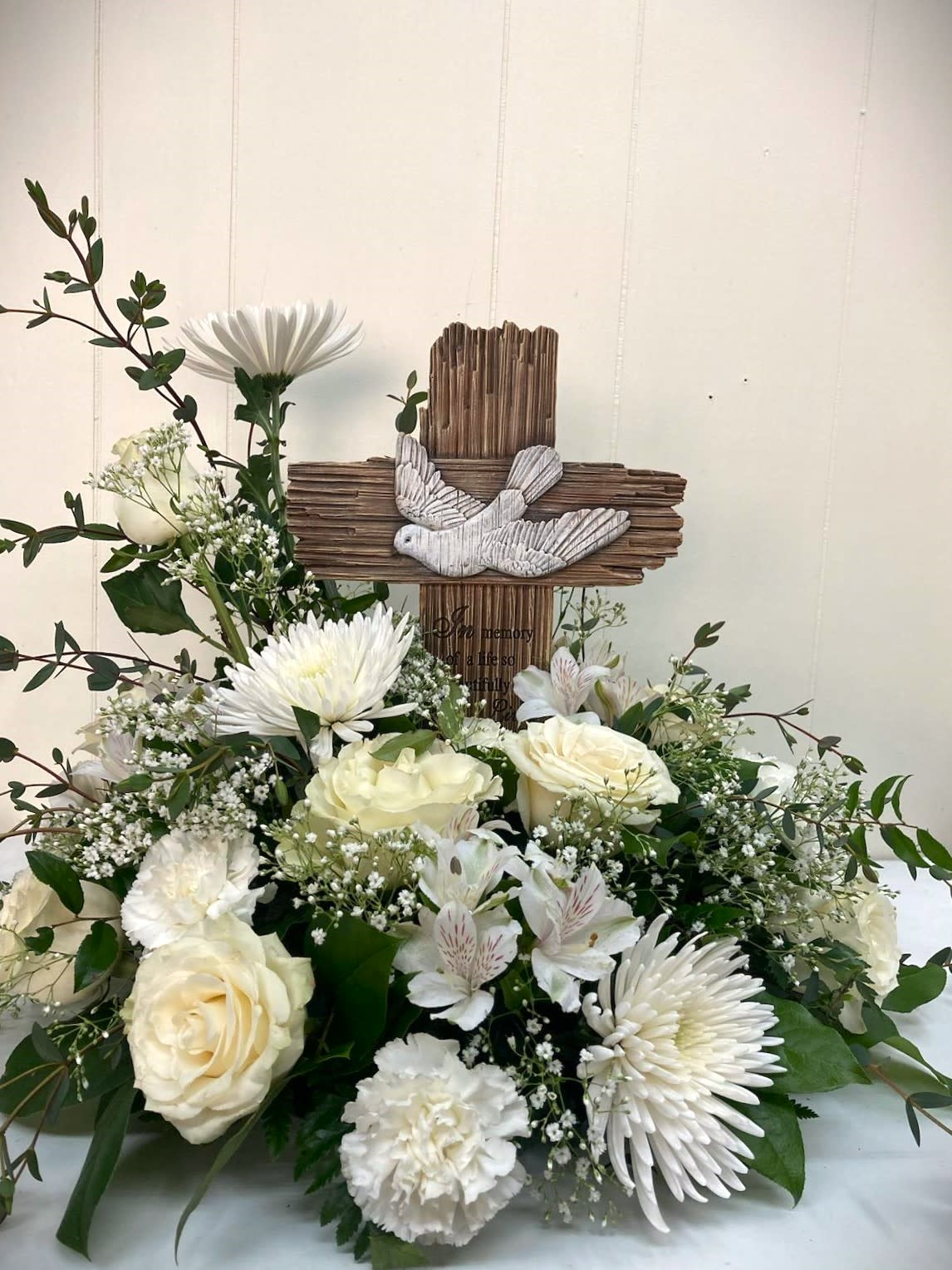 White floral arrangement with roses and daisies around a wooden cross