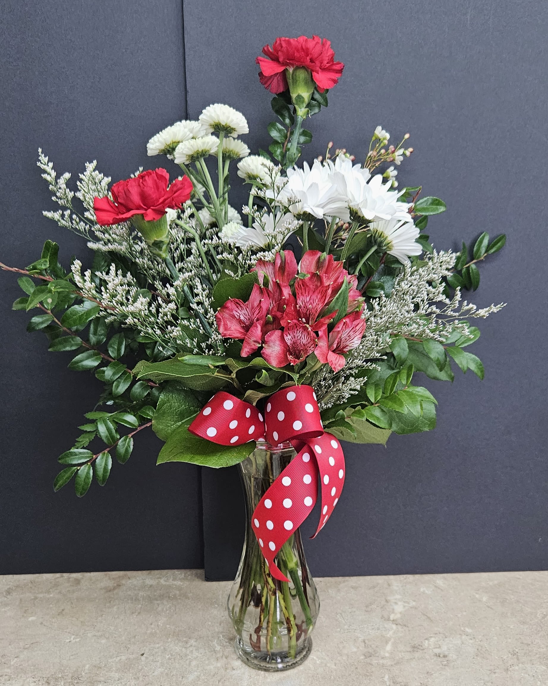 Bouquet of red carnations, white daisies, and pink alstroemeria in a glass vase with a red polka-dot ribbon.