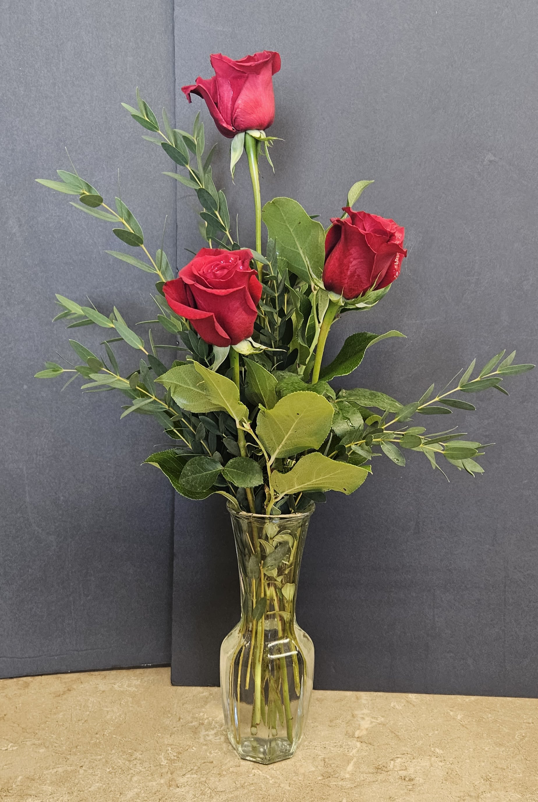 Three red roses in a clear glass vase with greenery