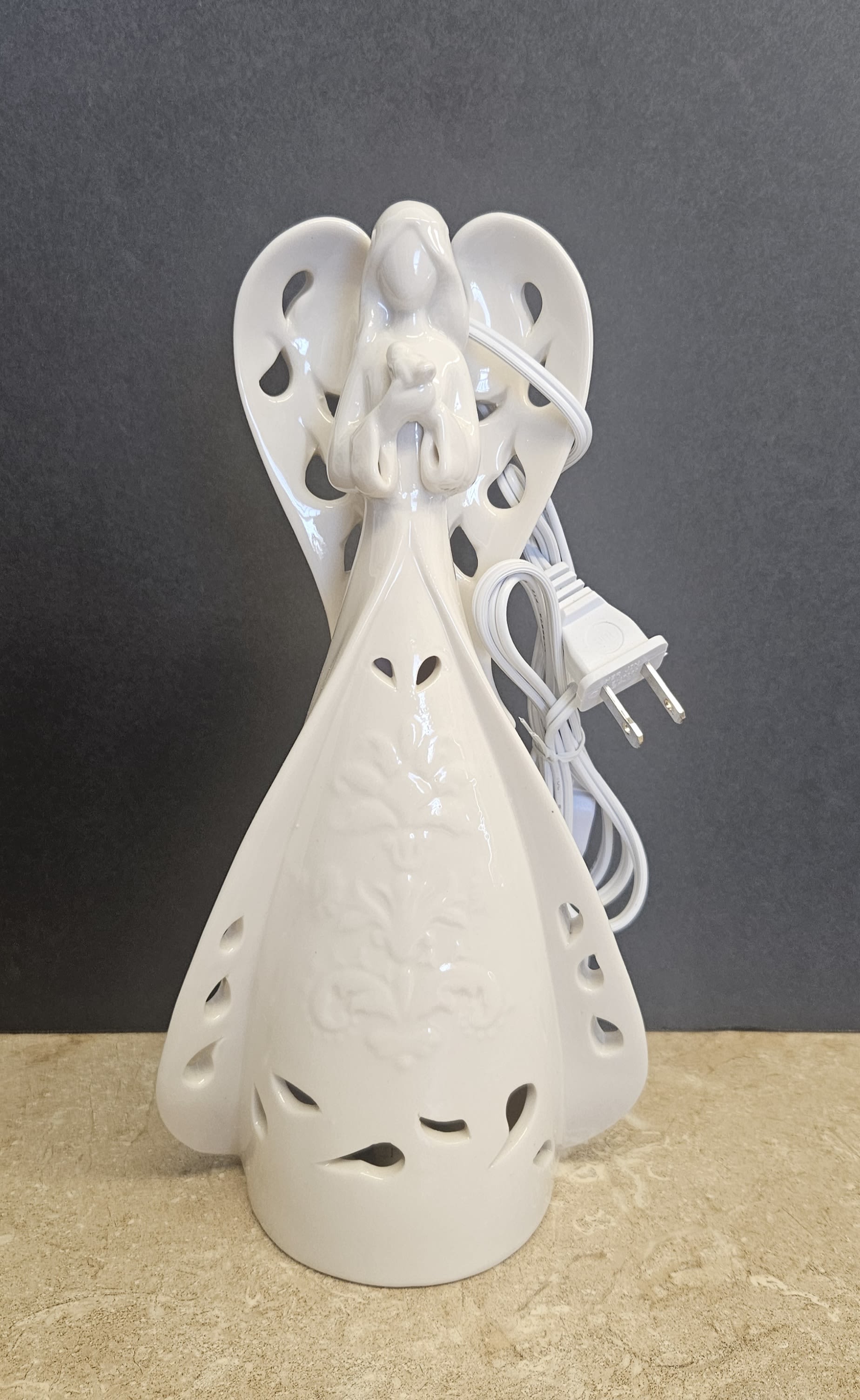 Ceramic Angel of light 12" - A softly glowing ceramic angel offers quiet comfort and gentle illumination - perfect as a sympathy or memorial gift or a serene accent for a nursery or Mother's Day mantel. This 12" white angel arrangement reads as a devotional keepsake rather than a bouquet, making it an especially thoughtful choice for New Baby blessings, funerals, or any time you want to send a message of peace and reassurance to the person you're thinking of.  Handcrafted in glossy white ceramic with delicate cutout details and outstretched wings, the angel stands 10" tall atop a classic gown silhouette. The piece includes a small square glass candle holder and a lighted element with a white power cord and two-prong plug for continuous soft illumination. The overall palette is pure white with neutral tones, so it complements nursery décor, memorial tables, or quiet corners of the home without competing with other décor.  Give this angel when words fall short - it's the kind of gift that helps someone feel seen, comforted, and remembered. Easy to place and simple to operate, the lighted figurine arrives ready to plug in and fill a room with a warm, contemplative glow; ideal for sending sympathy, celebrating a new baby, or marking Mother's Day with a calm, enduring keepsake.