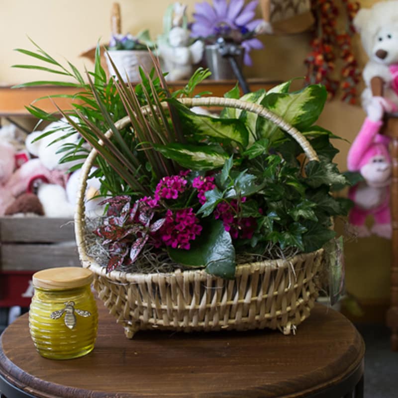 Basket arrangement with green foliage and pink flowers beside a yellow jar candle