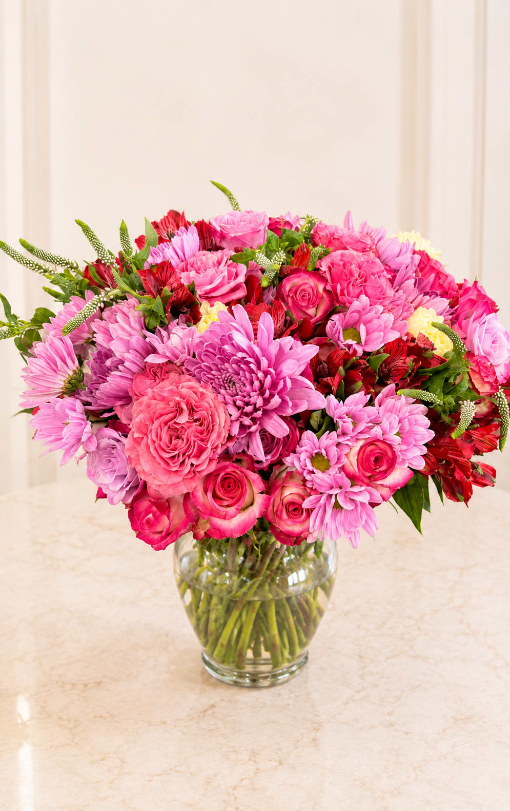 Pink Multicolored Bouquet- Designer Choice - A bright, full bouquet of mixed color blooms that reads as both joyful and romantic - perfect as a birthday centerpiece, a "thinking of you" surprise, or a just-because pick-me-up. This multicolored vase arrangement blends roses with pink and red Alstroemeria and chrysanthemums for a layered, lively look that still feels intimate enough for an anniversary or a love-and-romance moment.  Hand-delivered in a clear glass vase, the designer choice composition features carnations, roses, Alstroemeria, and chrysanthemums arranged into a rounded, abundant shape with slender green accent spikes for height and texture. Colors lean pink and purple with lively mixed color pops (photos show touches of red, white, and green), creating a cheerful contrast that brightens hospital rooms, new homes, or office desks. Vase dimensions and exact flower counts vary by size selected; each bouquet is professionally hydrated and arranged to arrive ready to display.  Choose this arrangement when you want the recipient to feel noticed and celebrated - a thoughtful option for get well, thank you, new baby, housewarming, Mother's Day, or just because. Available for same-day delivery around Houston when ordered early; stems arrive fresh with simple care instructions so flowers stay vibrant through spring and summer occasions.
