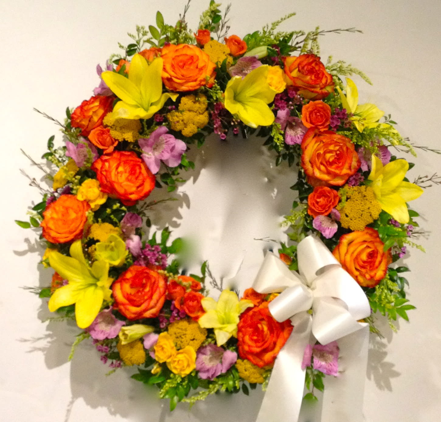 Circular floral wreath with orange roses, yellow lilies, and a white ribbon bow